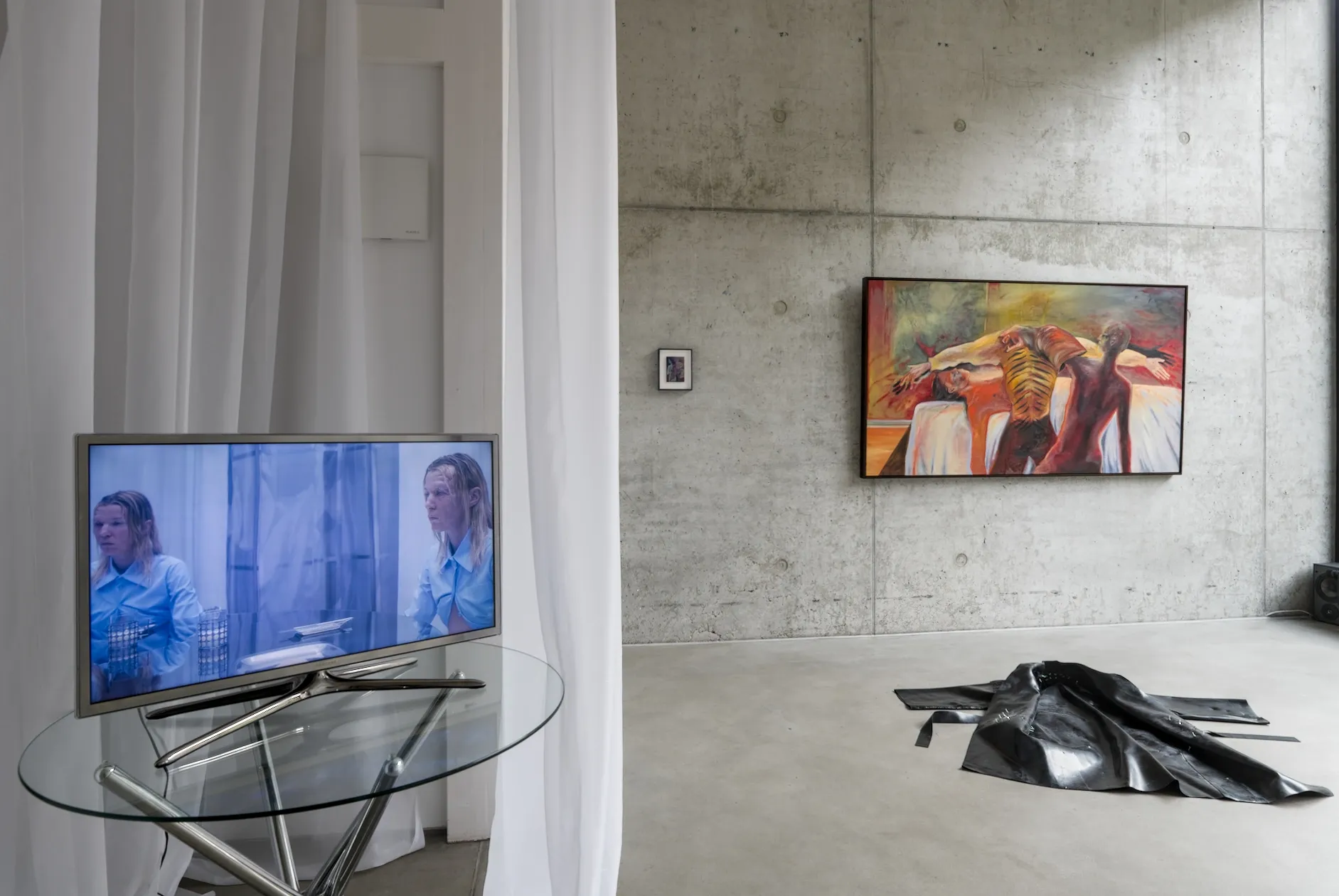 Exhibition review of I Think My Body No Longer Belongs to Me at roam projects e. V., Berlin, 2026. Monitor on glass table in front, painting and coat behind.