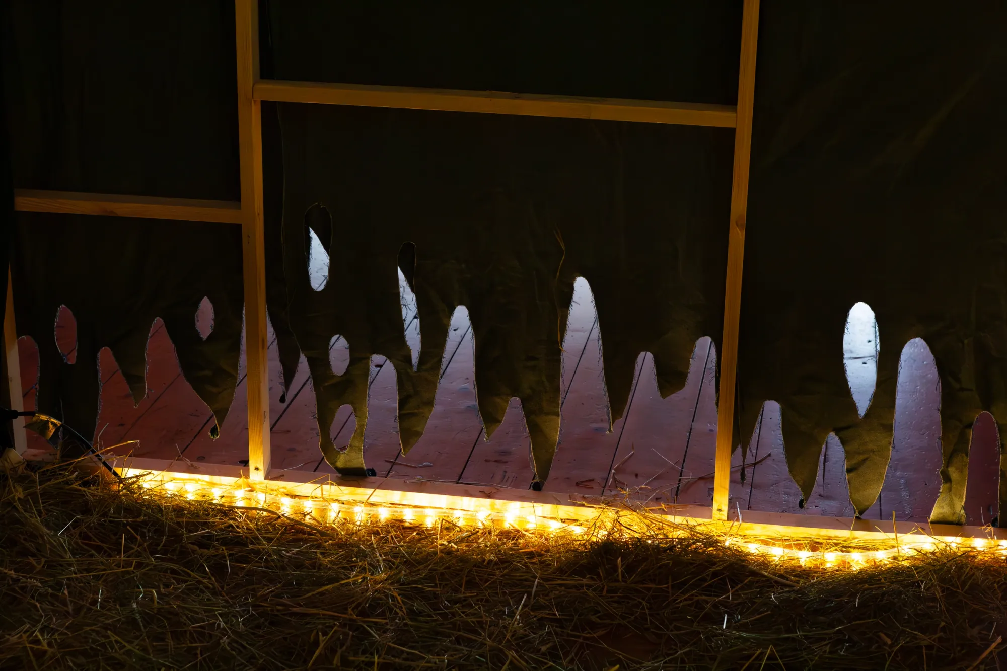 Exhibition review of Josefin Arnell: A Charm of Powerful Trouble at ACUD Galerie, Berlin, 2026. Darkened room lined with hay and warm fairy lights, video projected across the far wall showing two women outdoors.