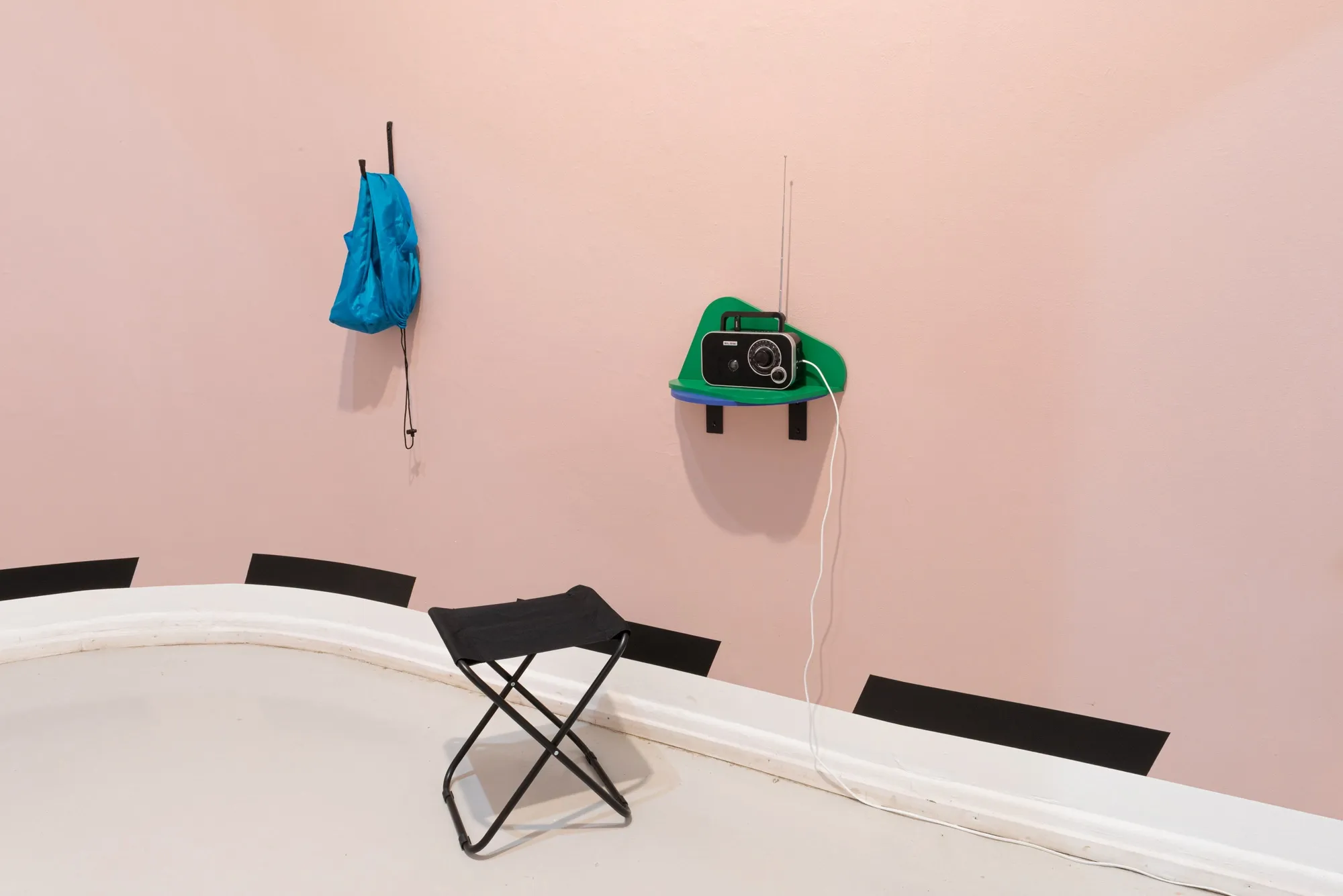 Exhibition review of Kaija Hinkula: You Are Here at Ama Gallery, Helsinki, 2026. A blue drawstring bag on a hook beside a green shelf holding a radio.