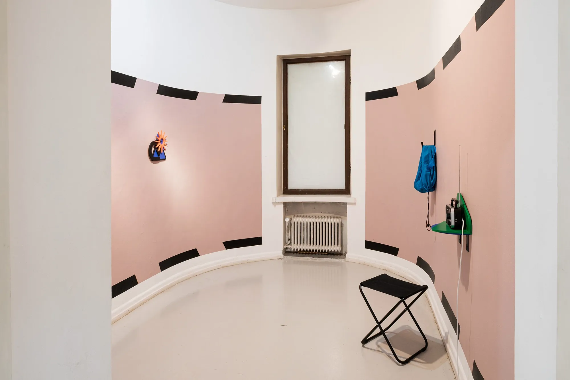 Exhibition review of Kaija Hinkula: You Are Here at Ama Gallery, Helsinki, 2026. A curved pink room with a folding stool, a hanging bag and a shelf radio.