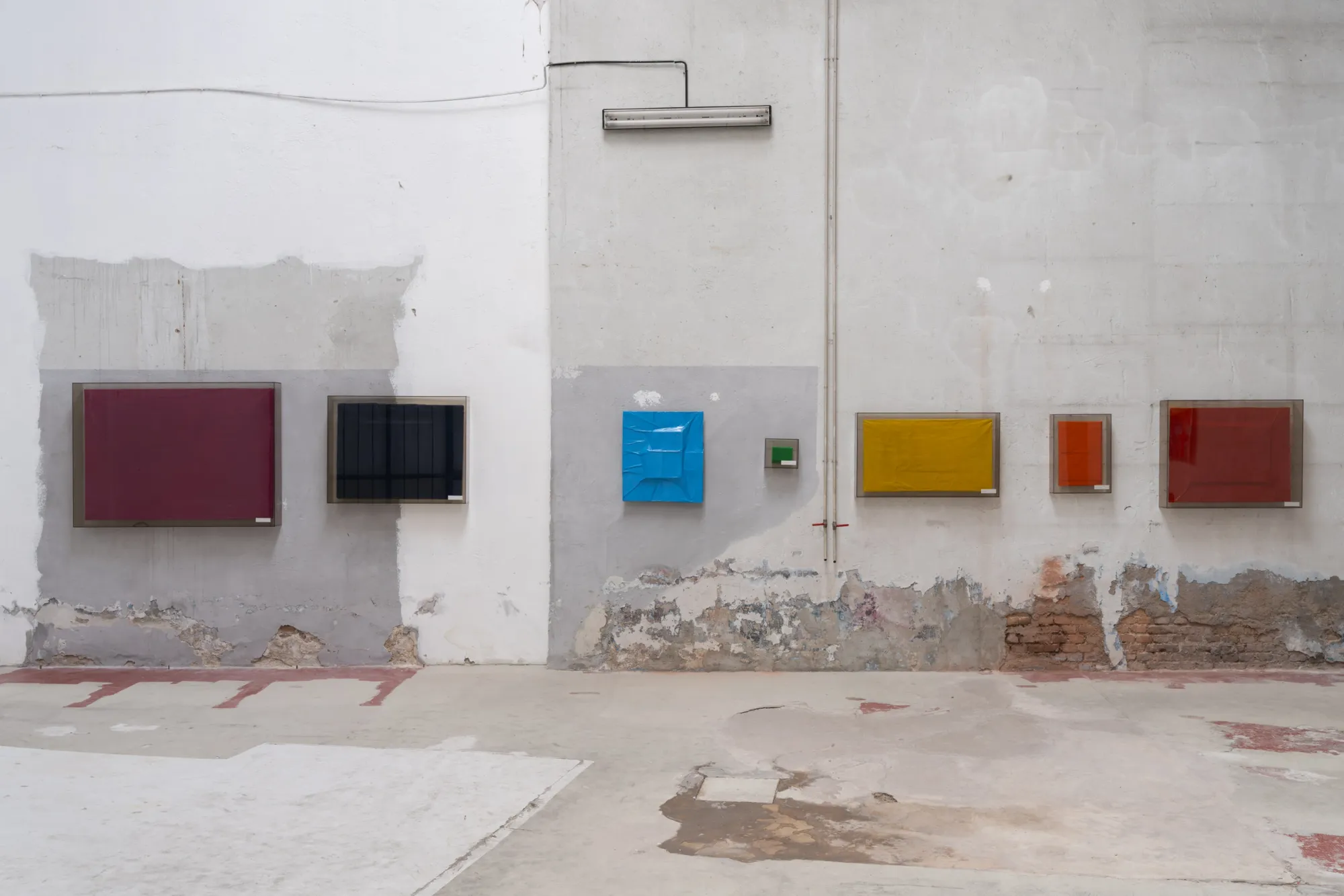 Exhibition review of Tillmann Lauterbach and Weseeclearly: Field Work at Factory Eleven, Milan, 2026. Row of framed monochrome panels in maroon, black, blue, yellow and red on a weathered, patched wall.