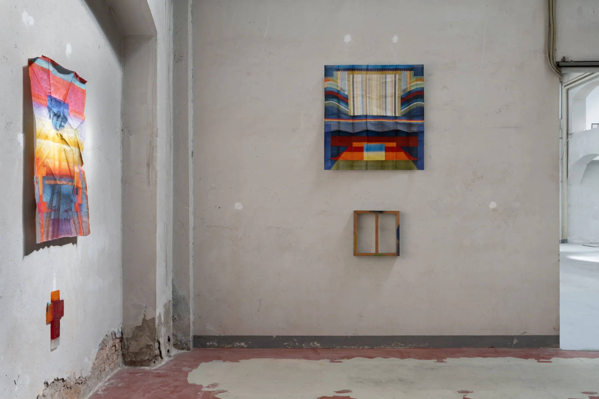 Exhibition review of Tillmann Lauterbach and Weseeclearly: Field Work at Factory Eleven, Milan, 2026. Worn industrial room with a folded textile and a red cross on one wall and framed panels on anothe