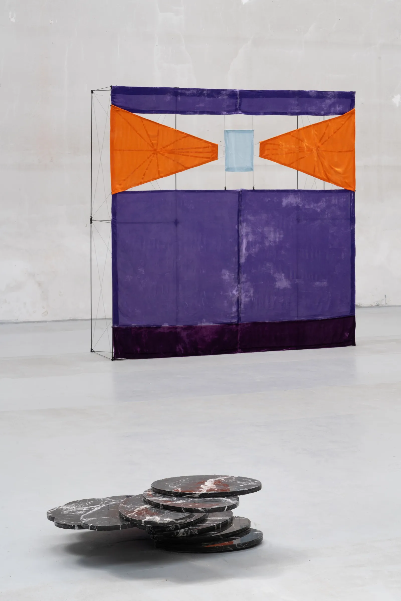 Exhibition review of Tillmann Lauterbach and Weseeclearly: Field Work at Factory Eleven, Milan, 2026. Violet dyed fabric screen with orange triangles on a thin frame, a stack of dark marble discs on the floor.