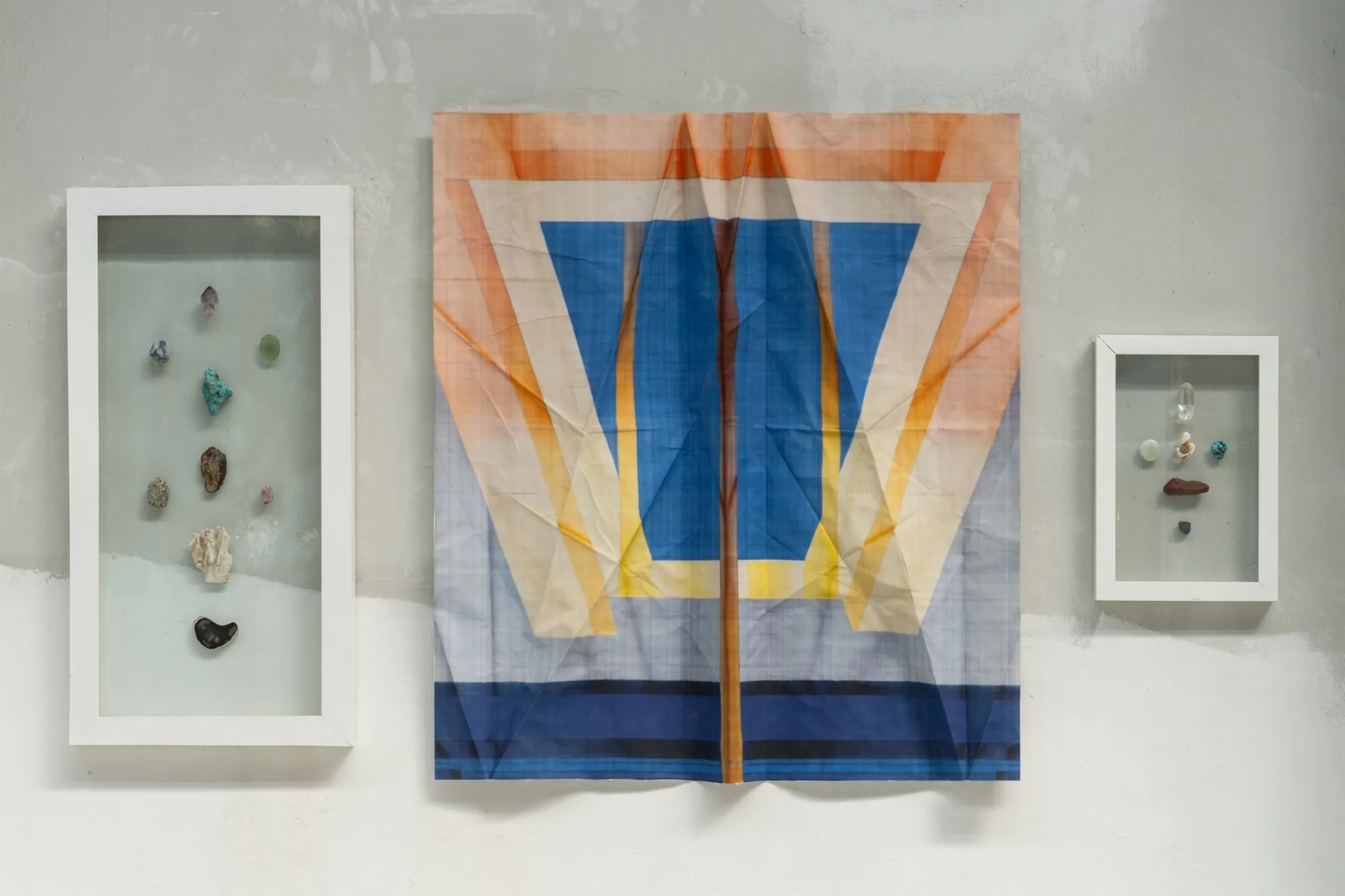 Exhibition review of Tillmann Lauterbach and Weseeclearly: Field Work at Factory Eleven, Milan, 2026. Folded textile with a blue and orange trapezoid between two white vitrines holding small stones.