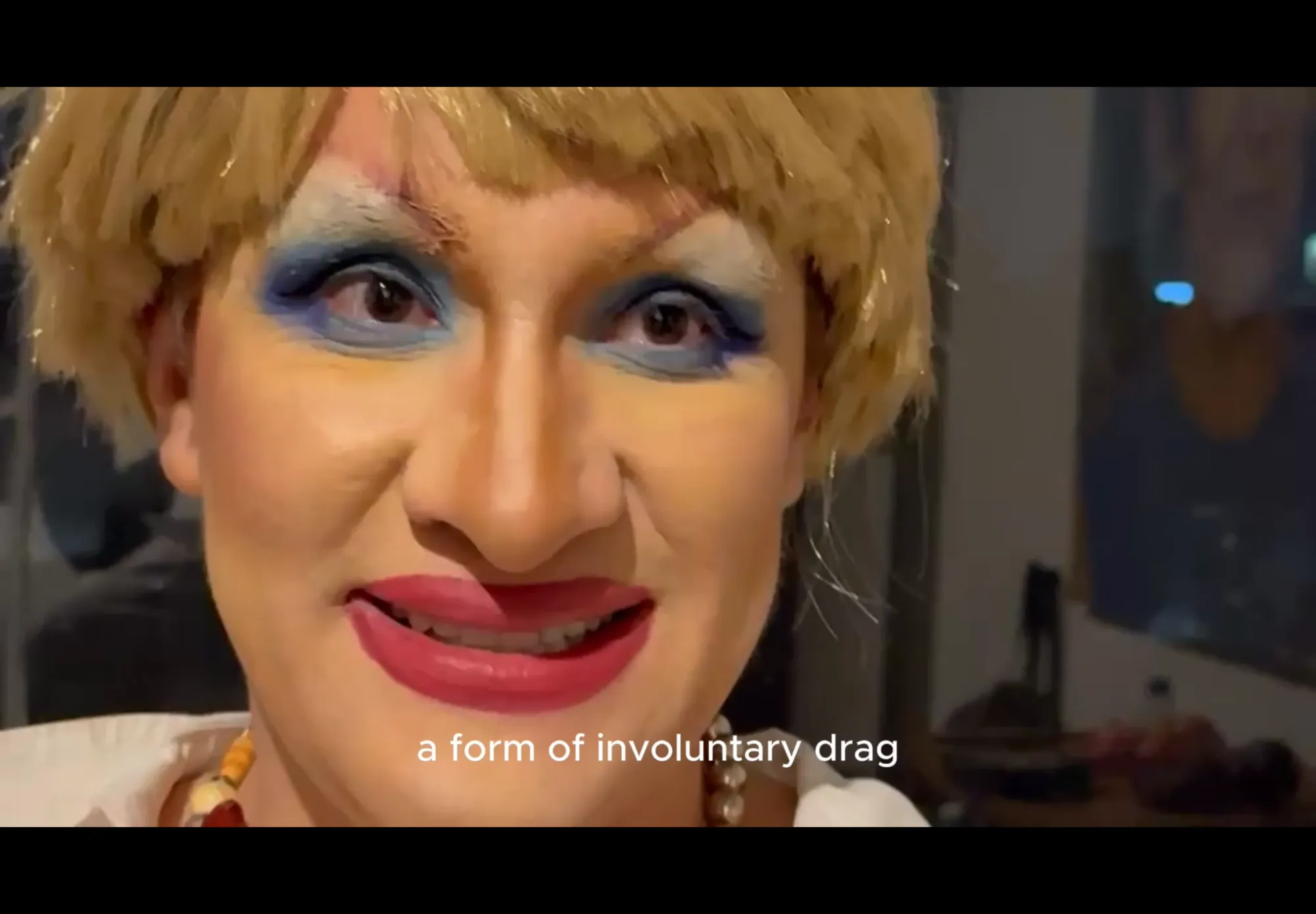 Exhibition review of Lic. Sniffany: A Charm of Powerful Trouble at ACUD Galerie, Berlin, 2026. Close-up video still of a face in exaggerated drag makeup with blonde wig and red lipstick.