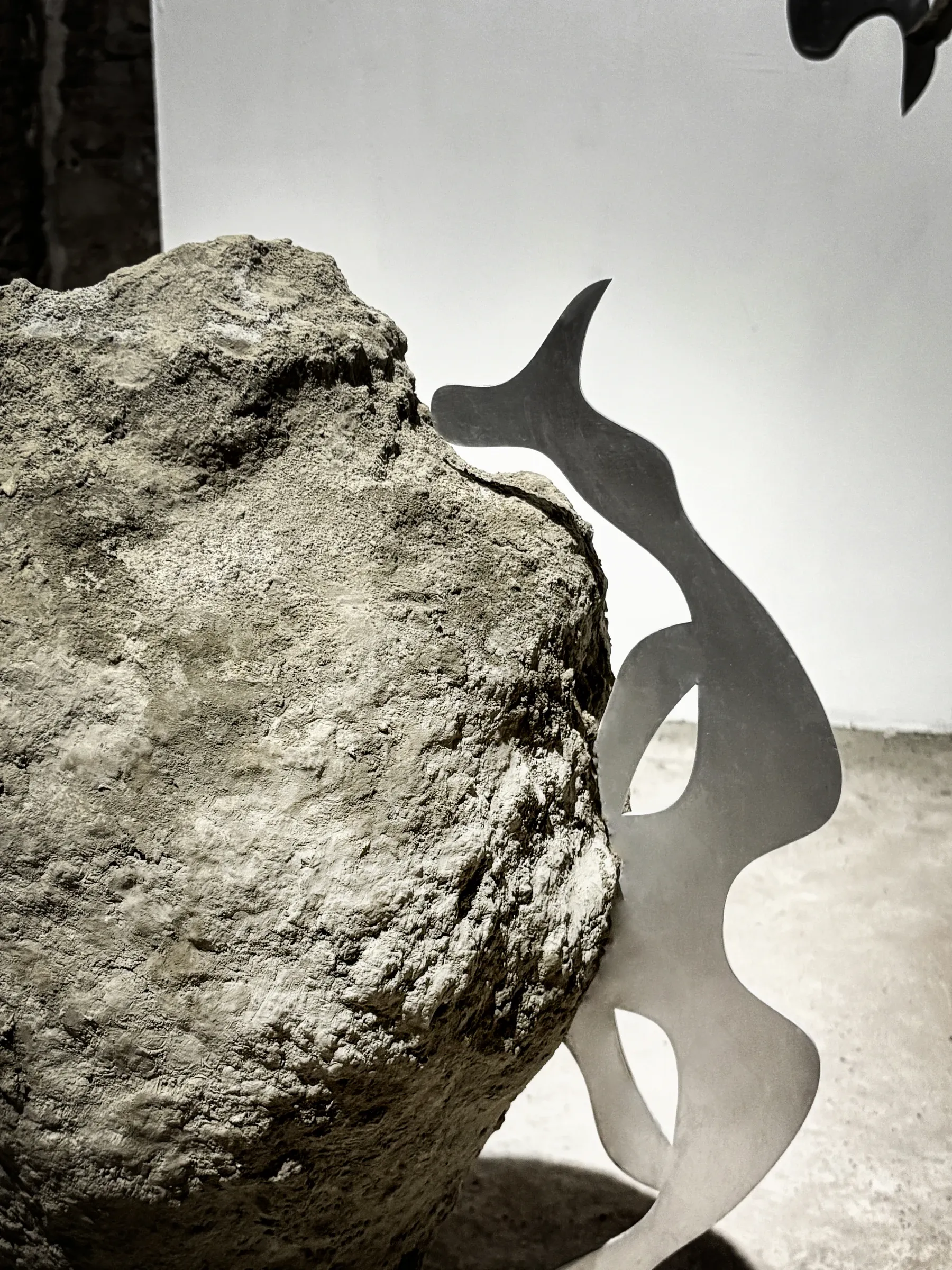 Exhibition review of Niki Danai Chania: Lamiae at the Agora, Bonifacio, 2026. Side view of a boulder with a single curved mirror-polished steel fin rising from a crevice against a plain wall.