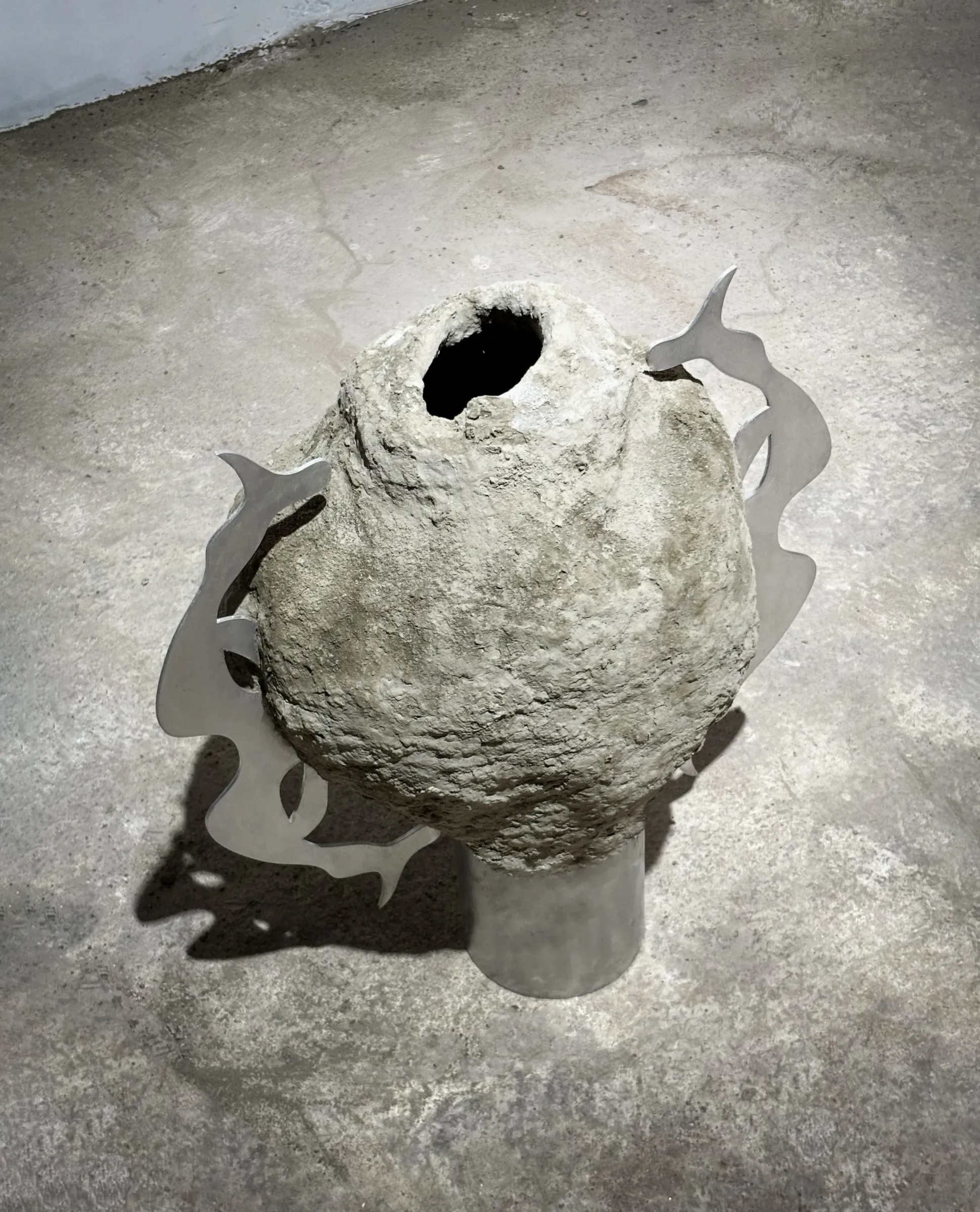 Exhibition review of Niki Danai Chania: Lamiae at the Agora, Bonifacio, 2026. Egg-shaped hollow concrete form with a dark opening at its crown, on a steel base flanked by curved steel fin shapes.