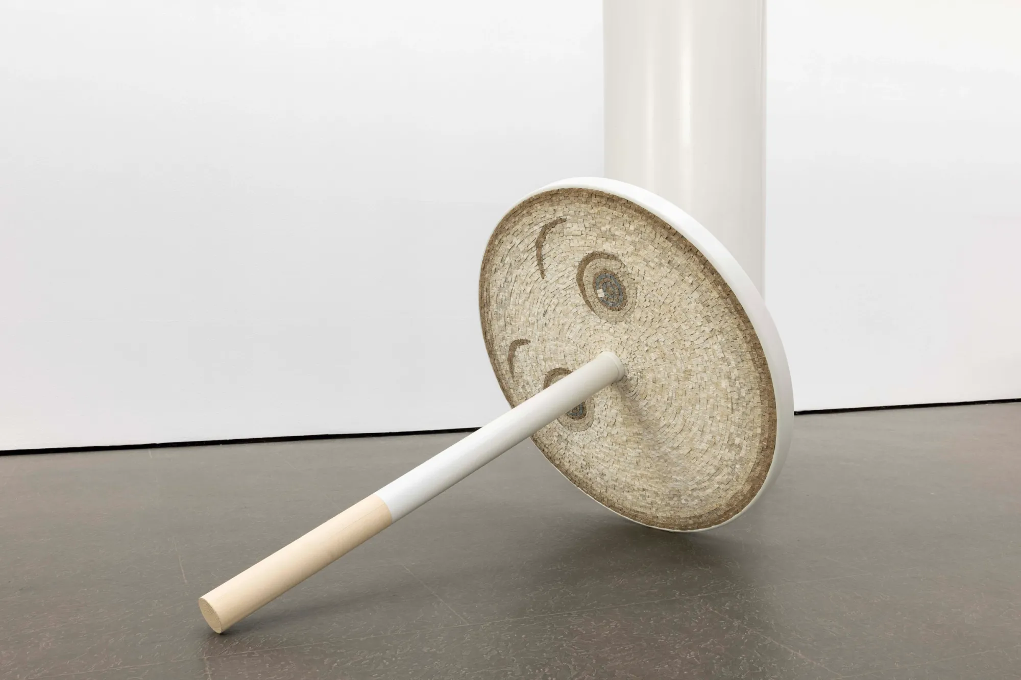 Exhibition review of Riikka Anttonen: Vertere at Sinne, Helsinki, 2026. Spinning-top sculpture leaning against a column, its disc showing a cream marble mosaic face with closed eyes.
