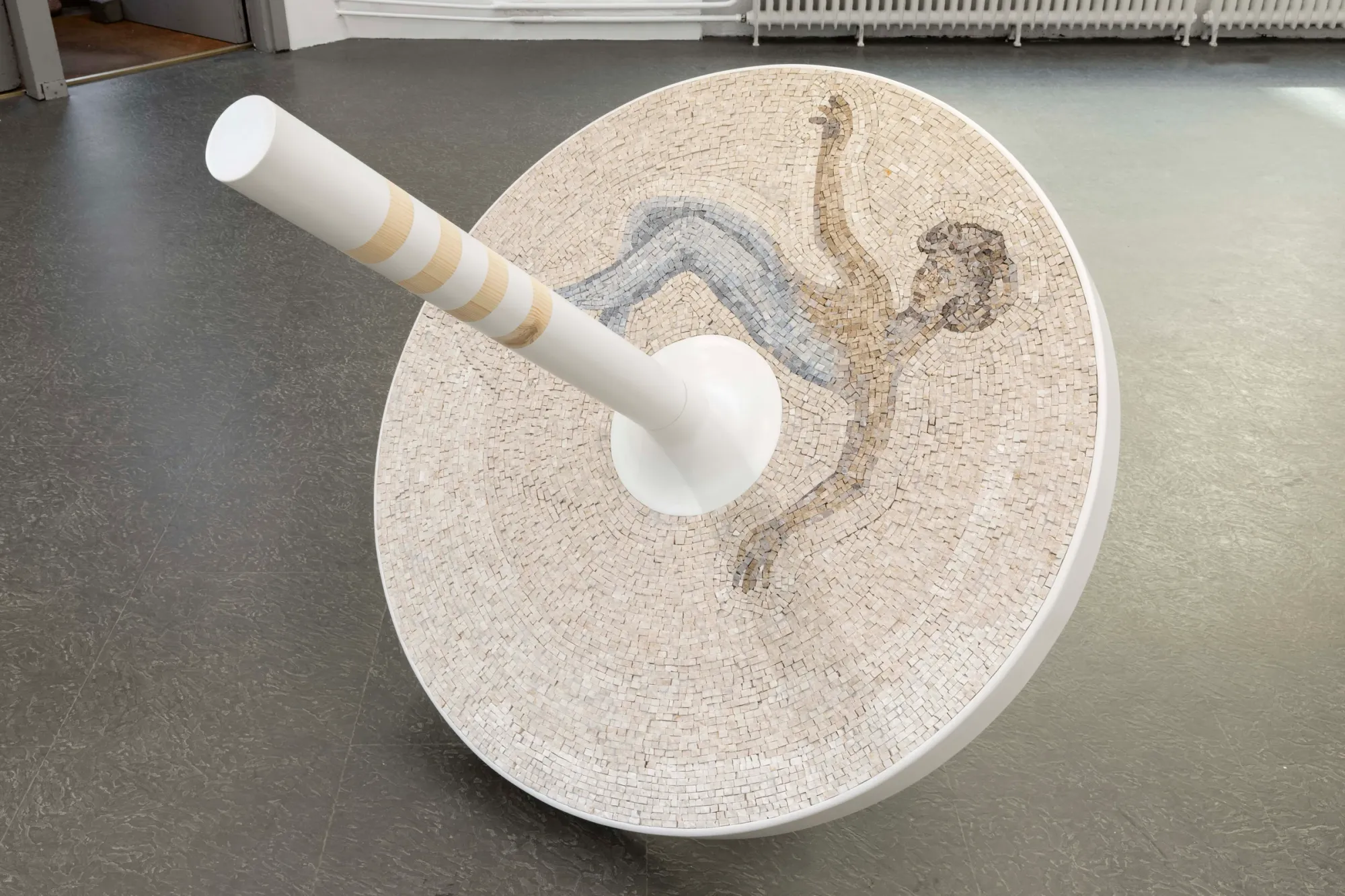 Exhibition review of Riikka Anttonen: Vertere at Sinne, Helsinki, 2026. Overhead view of a spinning-top sculpture with a marble mosaic figure caught mid-fall across its disc.