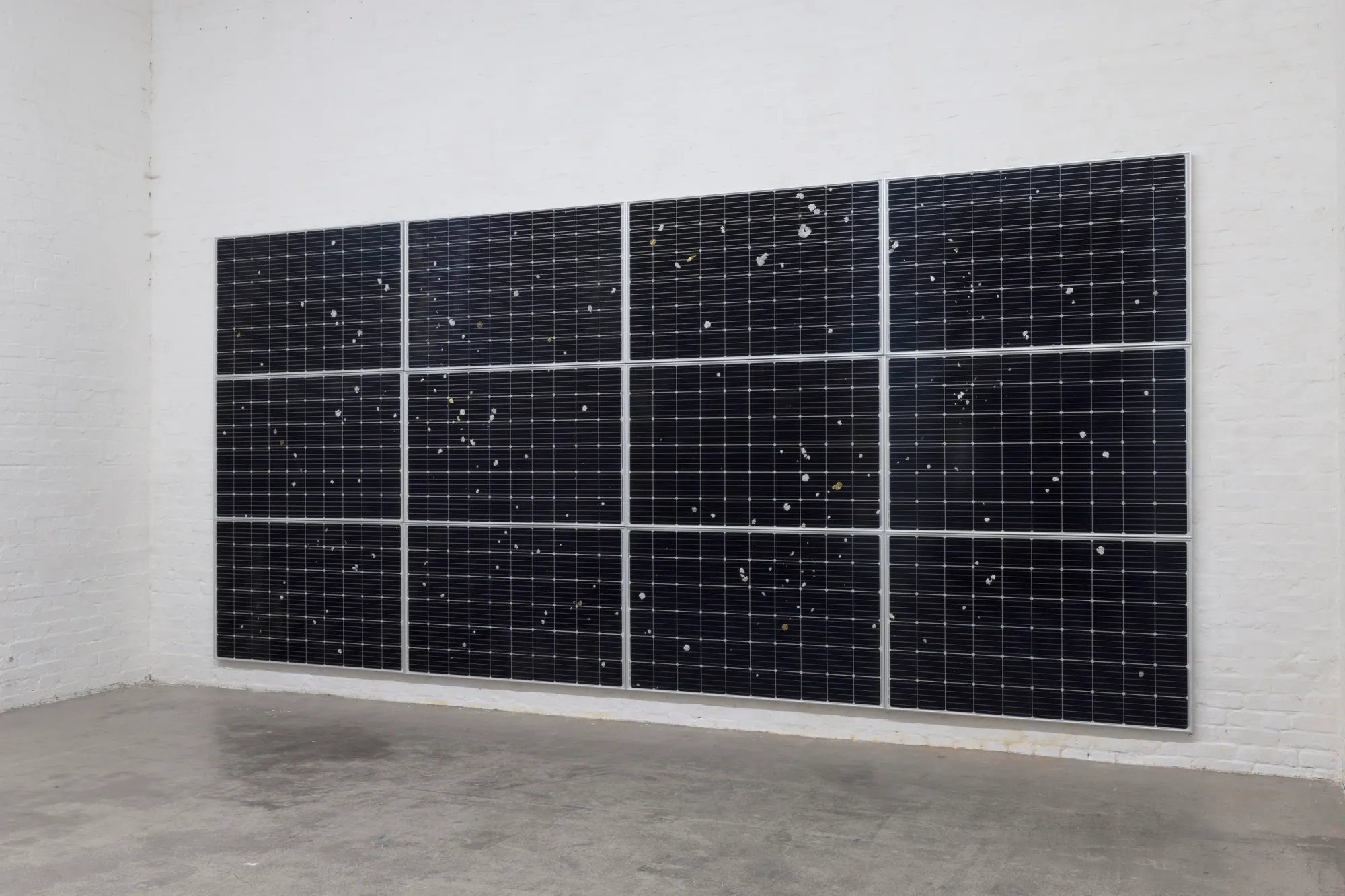 Exhibition review of Sari Palosaari: Substrate at Forum Box, Helsinki, 2026. Twelve black solar panels mounted as a wall grid, scattered with small silver and gold metal casts.