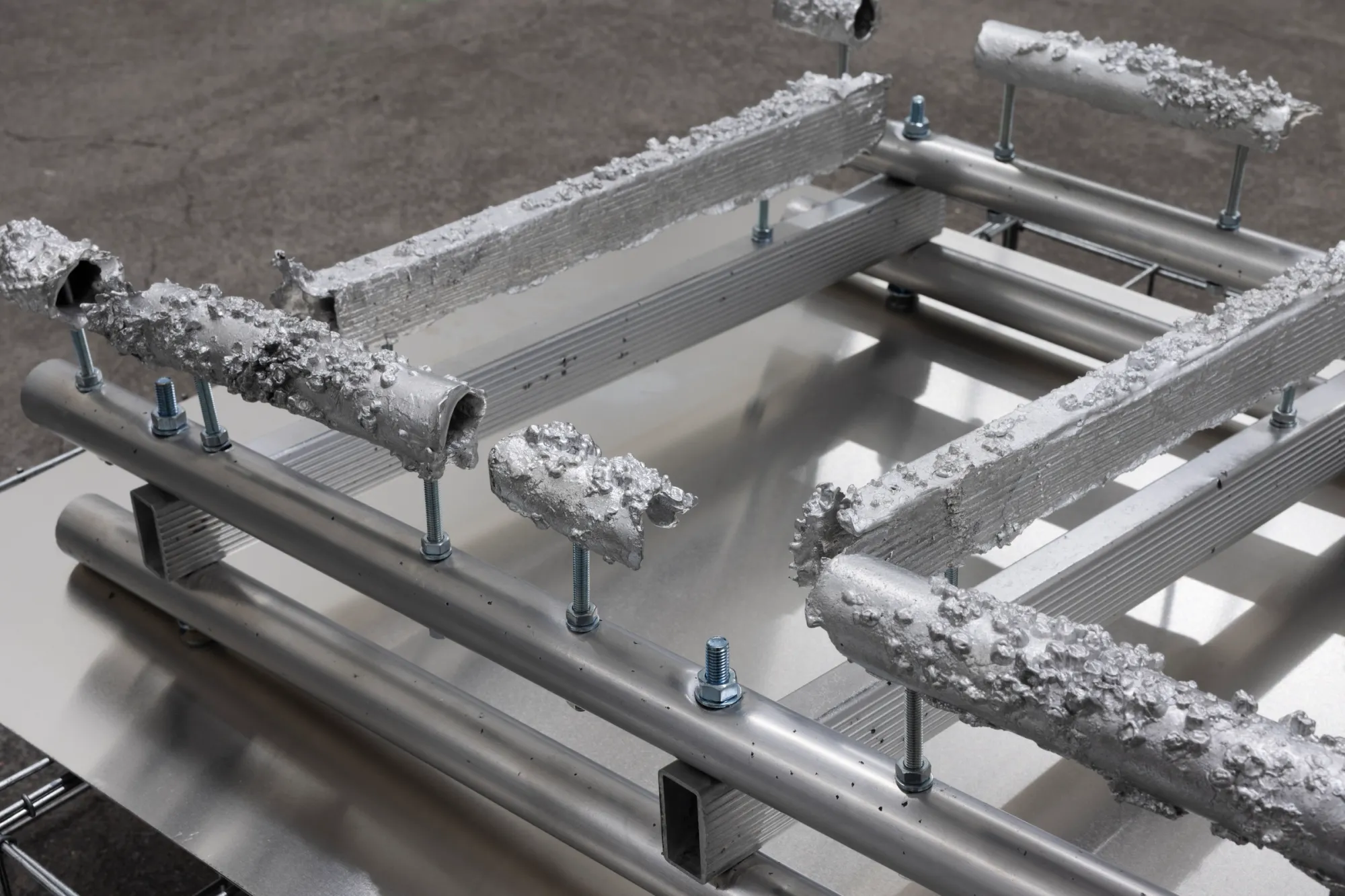Exhibition review of Sari Palosaari: Substrate at Forum Box, Helsinki, 2026. Aluminium tubes coated in rough cast lichen-like crusts, bolted onto a metal frame.
