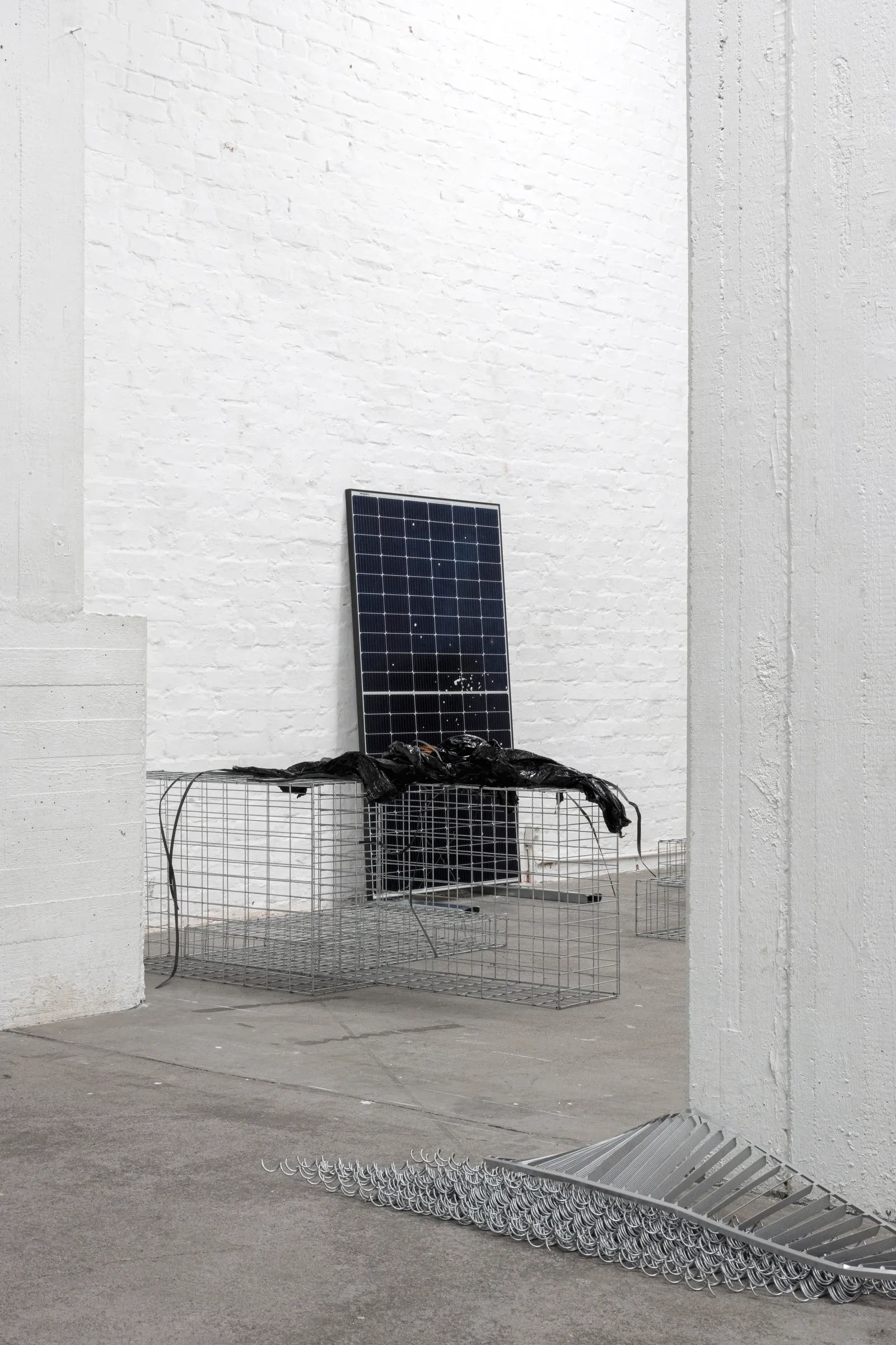 Exhibition review of Sari Palosaari: Substrate at Forum Box, Helsinki, 2026. Solar panel leaning on a wire gabion basket draped with black material beside a white pillar, metal parts on the floor.