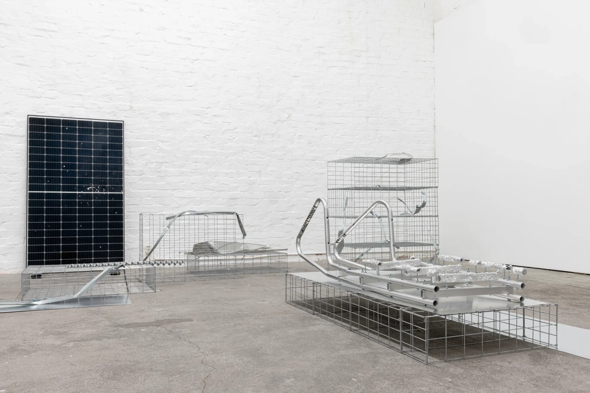 Exhibition review of Sari Palosaari: Substrate at Forum Box, Helsinki, 2026. White brick room with an upright solar panel, wire mesh gabion baskets and bent aluminium tubes on a concrete floor.