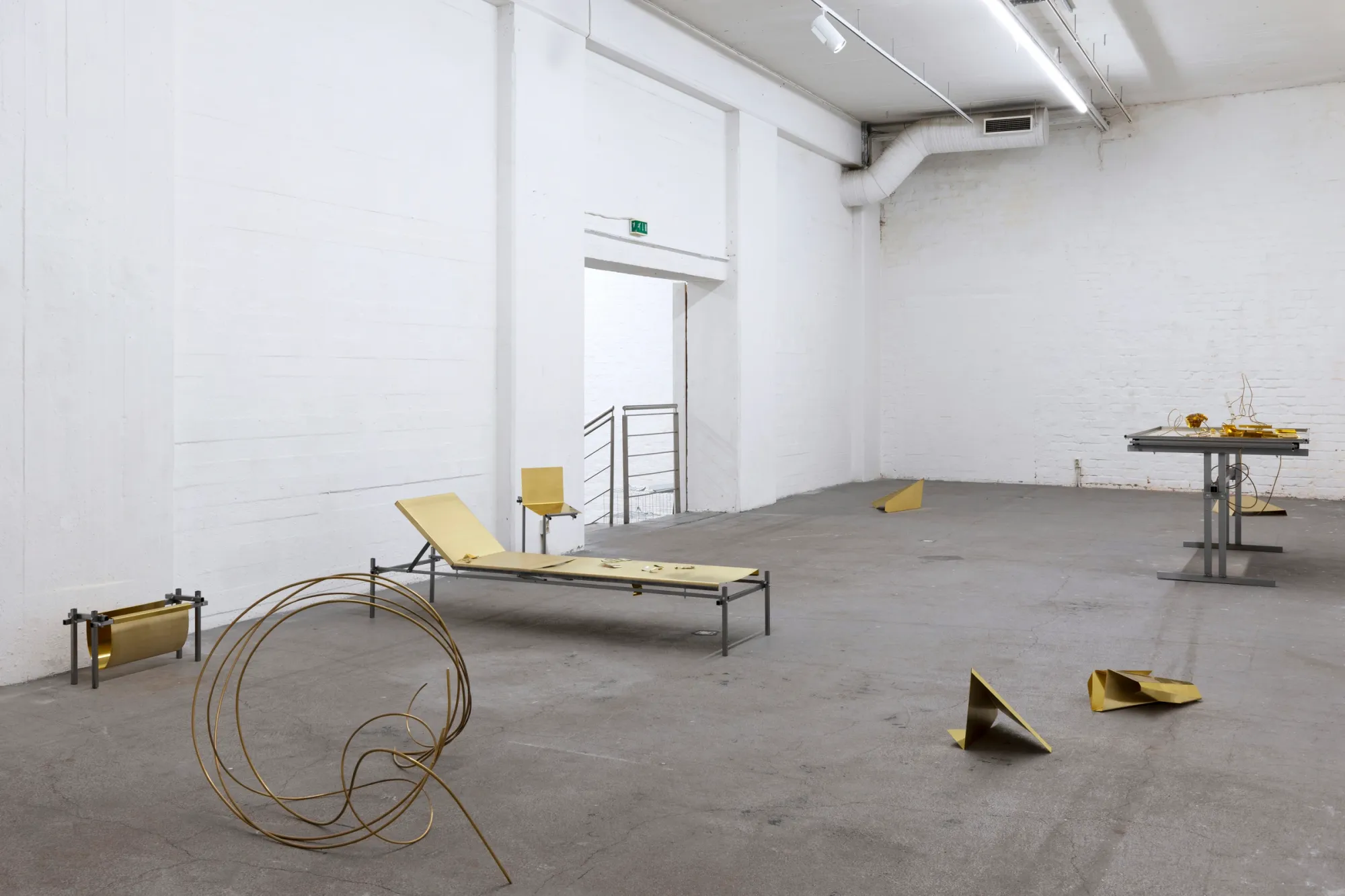 Exhibition review of Sari Palosaari: Substrate at Forum Box, Helsinki, 2026. Brass-topped low bench, a looped brass wire, folded brass pieces on the floor and a steel desk in the distance.