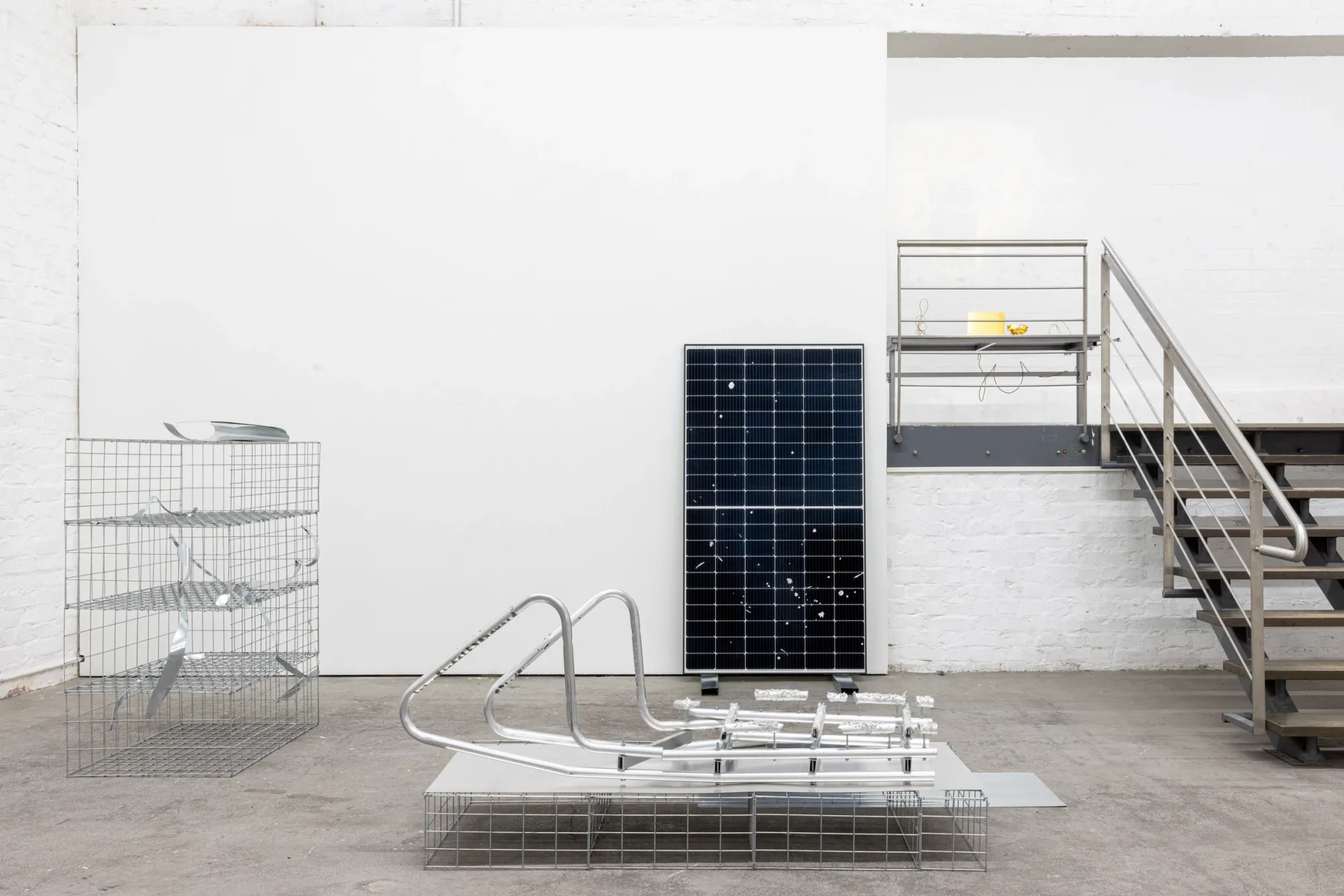 Exhibition review of Sari Palosaari: Substrate at Forum Box, Helsinki, 2026. Mesh gabion cage, bent aluminium tubes on a mesh base, an upright solar panel and a steel staircase to a mezzanine.