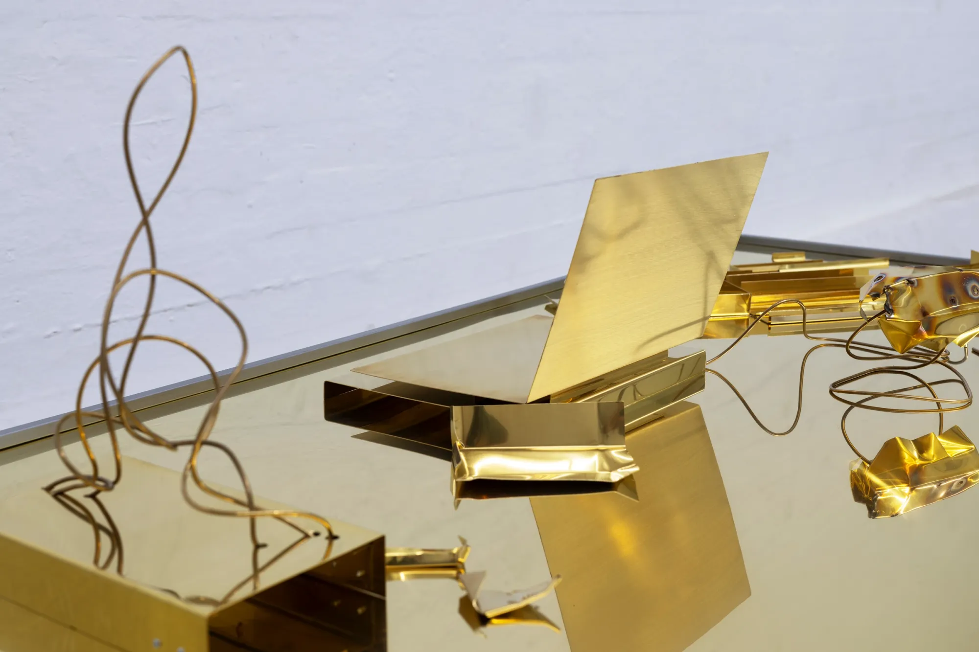 Exhibition review of Sari Palosaari: Substrate at Forum Box, Helsinki, 2026. Mirrored brass desktop with folded brass sheets, a curling brass wire and small crumpled brass forms.