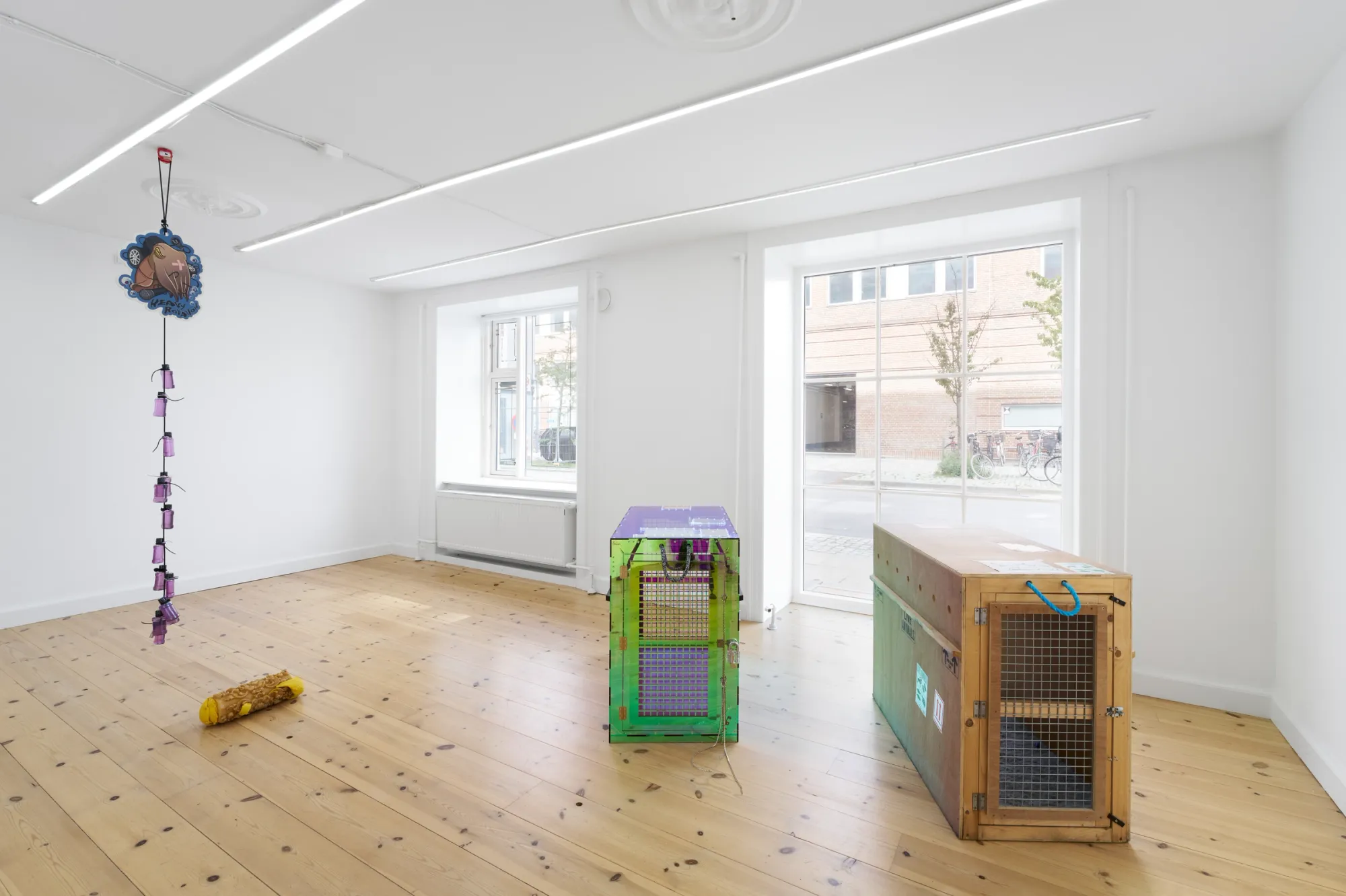 Exhibition review of Sequence for an Optimal Dispatch at aaaa nordhavn, Copenhagen, 2026. Hanging cord with pink bottles, an acrylic crate and a wooden crate by tall windows.