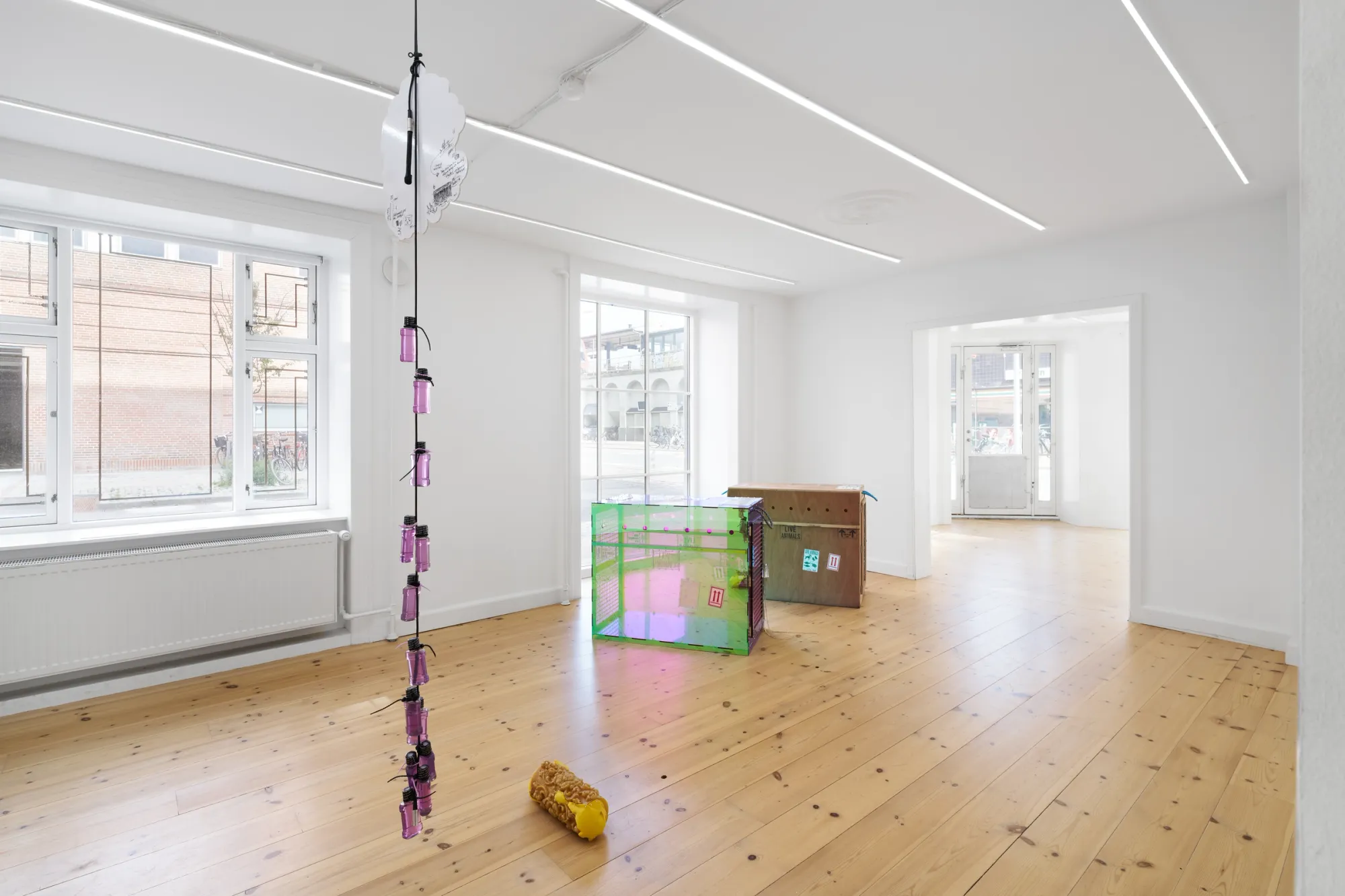 Exhibition review of Sequence for an Optimal Dispatch at aaaa nordhavn, Copenhagen, 2026. Pink bottles on a ceiling cord, yellow wax roll on the floor, two crates behind.