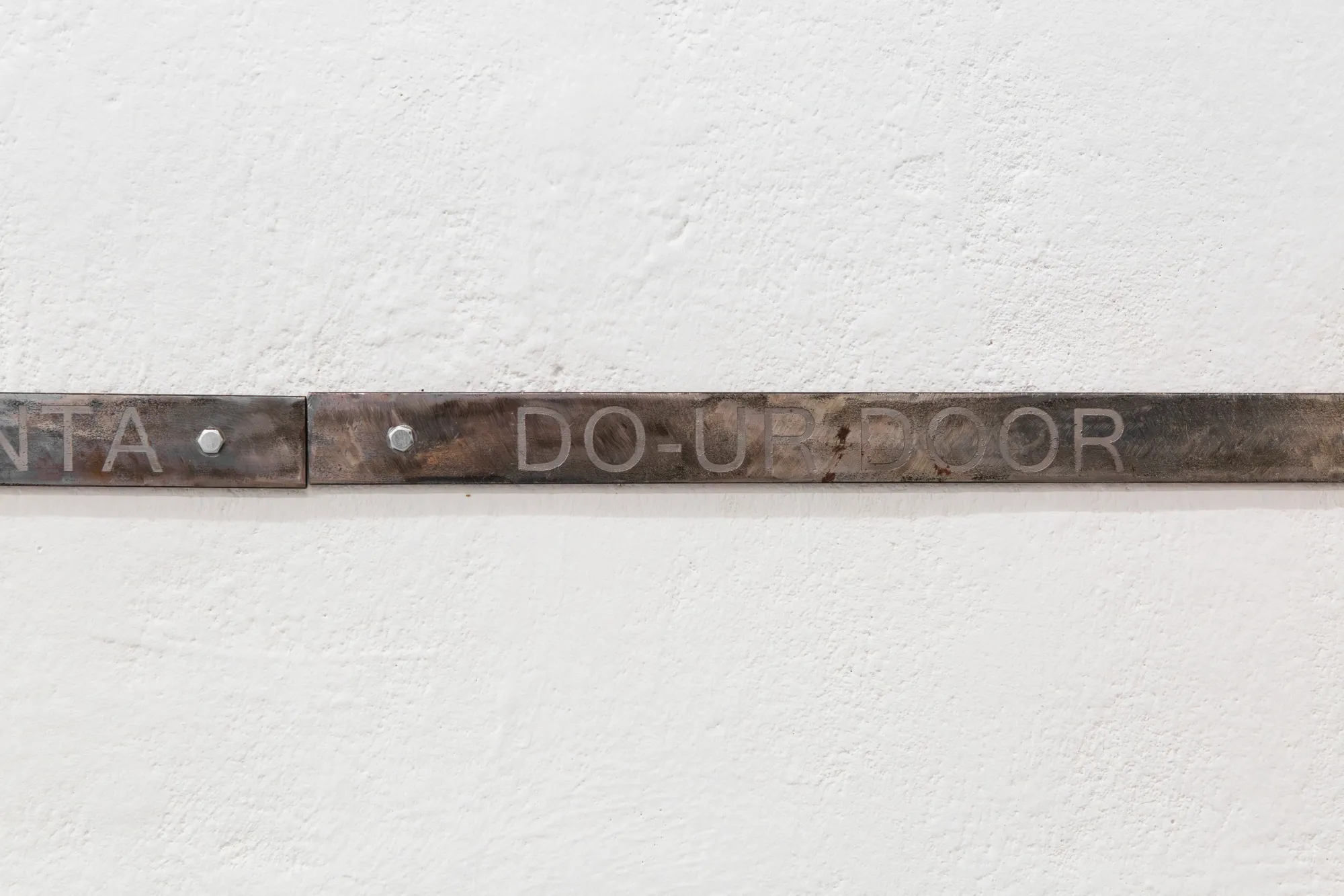 Exhibition review of TLC23: A Charm of Powerful Trouble at ACUD Galerie, Berlin, 2026. Close view of an etched metal bar mounted on a white wall reading DO-UP DOOR