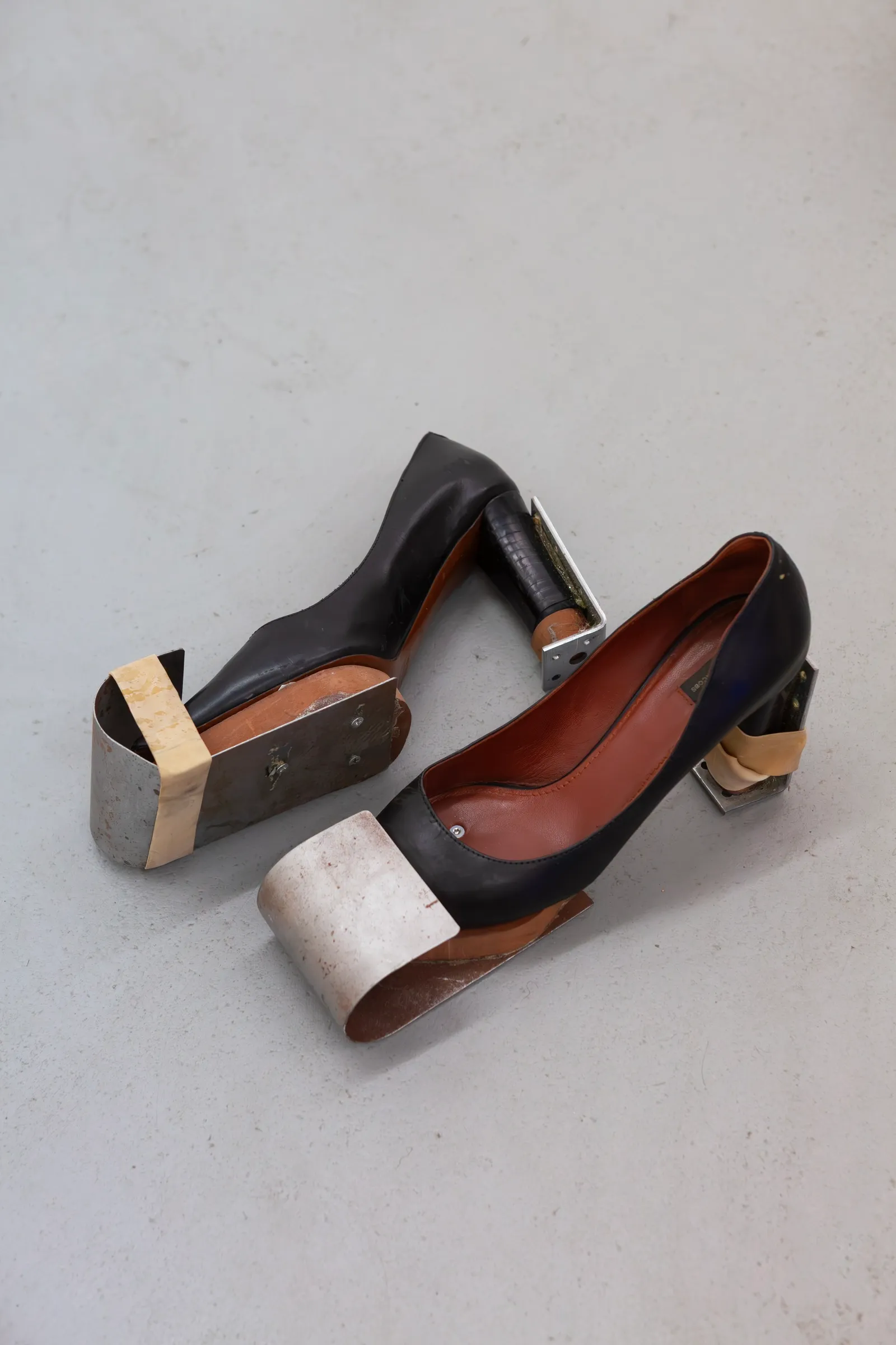 Exhibition review of TLC23: A Charm of Powerful Trouble at ACUD Galerie, Berlin, 2026. A pair of black leather high-heeled shoes wrapped with raw metal cuffs, placed directly on the concrete floor.