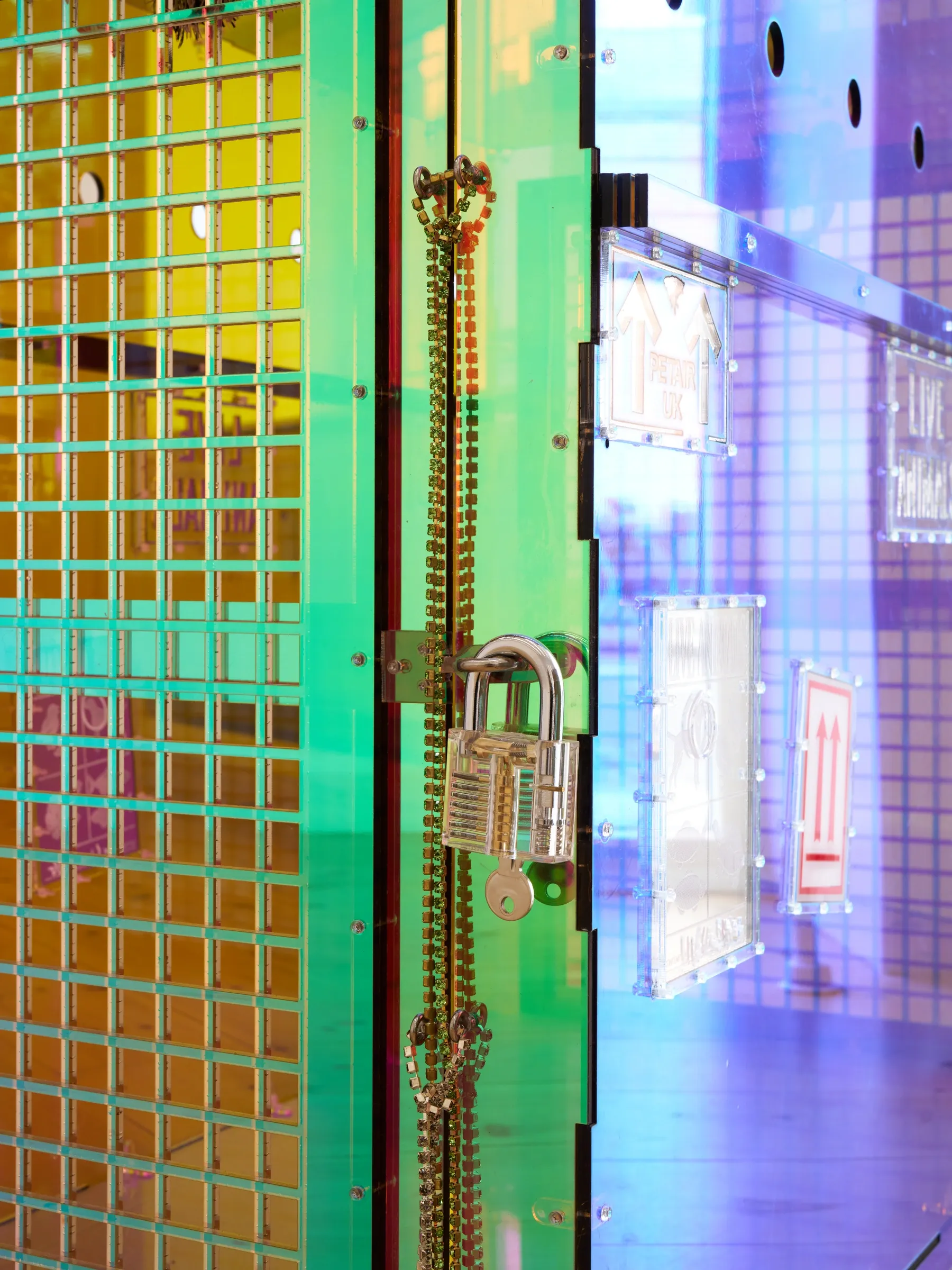 Exhibition review of Villiam Miklos Andersen: Sequence for an Optimal Dispatch at aaaa nordhavn, Copenhagen, 2026. Transparent padlock and chain on an iridescent green acrylic grid door.