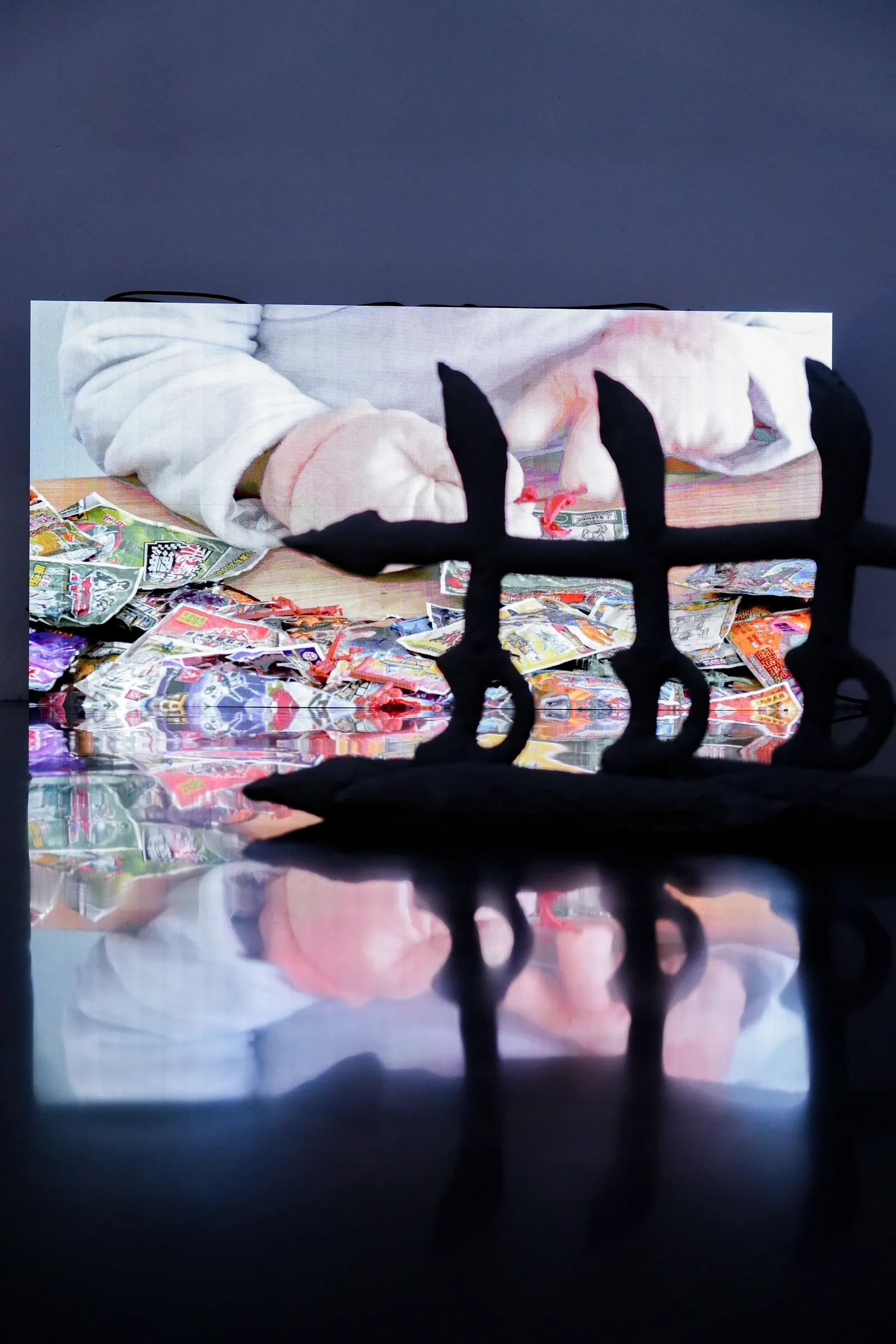 Exhibition review of Zhang Ting: TechDiumMory at TANK Shanghai, Shanghai, 2026. Video screen of hands over toy packets behind a black fence form on a reflective floor.