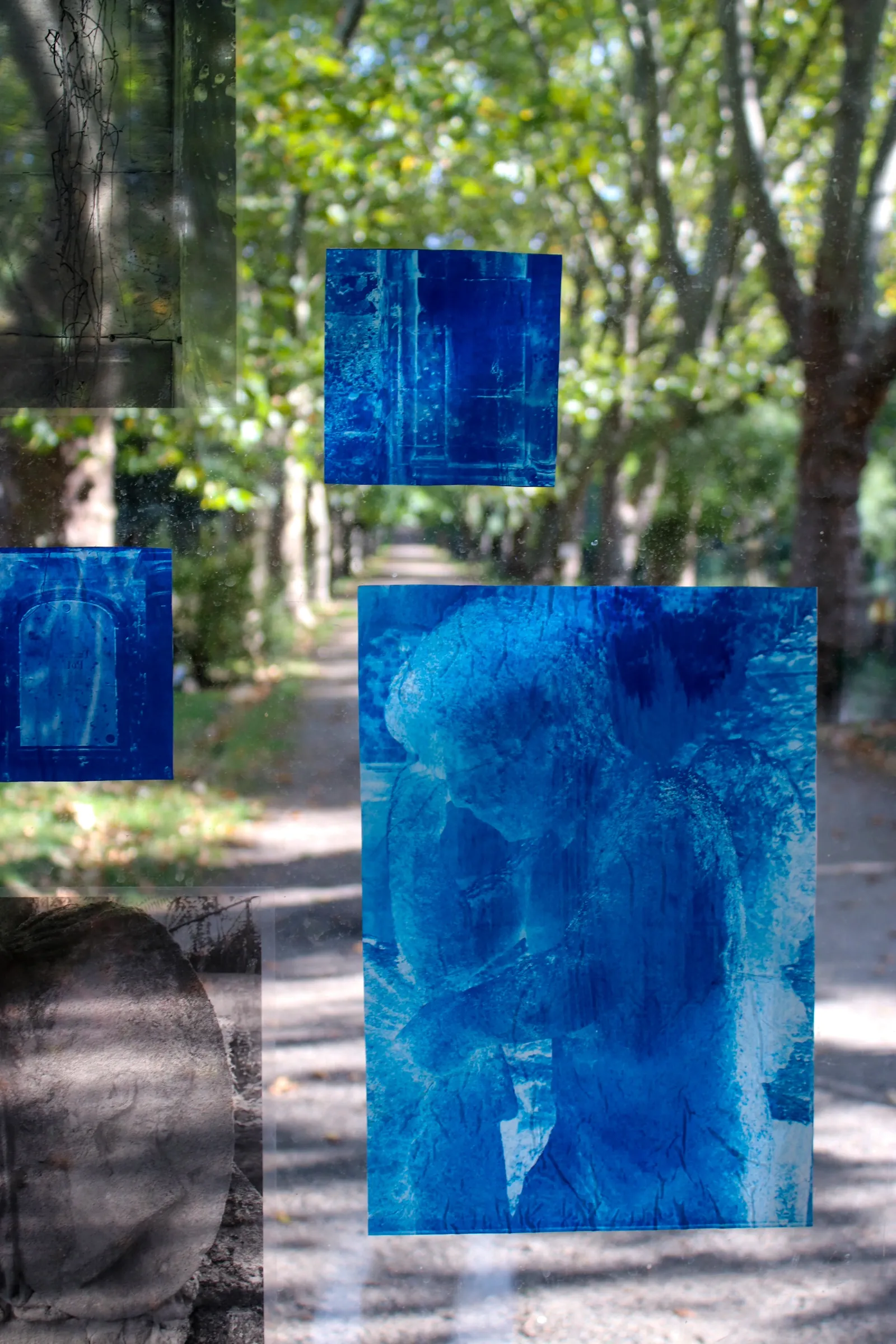 Aubrey&nbsp;Theobald&nbsp;and&nbsp;Kristina&nbsp;Popov,&nbsp;Phantom&nbsp;Pförtnerhaus&nbsp;Chapter&nbsp;3,&nbsp;Berlin,&nbsp;2026.&nbsp;Blue&nbsp;and&nbsp;grey&nbsp;photographic&nbsp;prints&nbsp;mounted&nbsp;on&nbsp;a&nbsp;window&nbsp;with&nbsp;trees&nbsp;and&nbsp;a&nbsp;cemetery&nbsp;path&nbsp;behind.