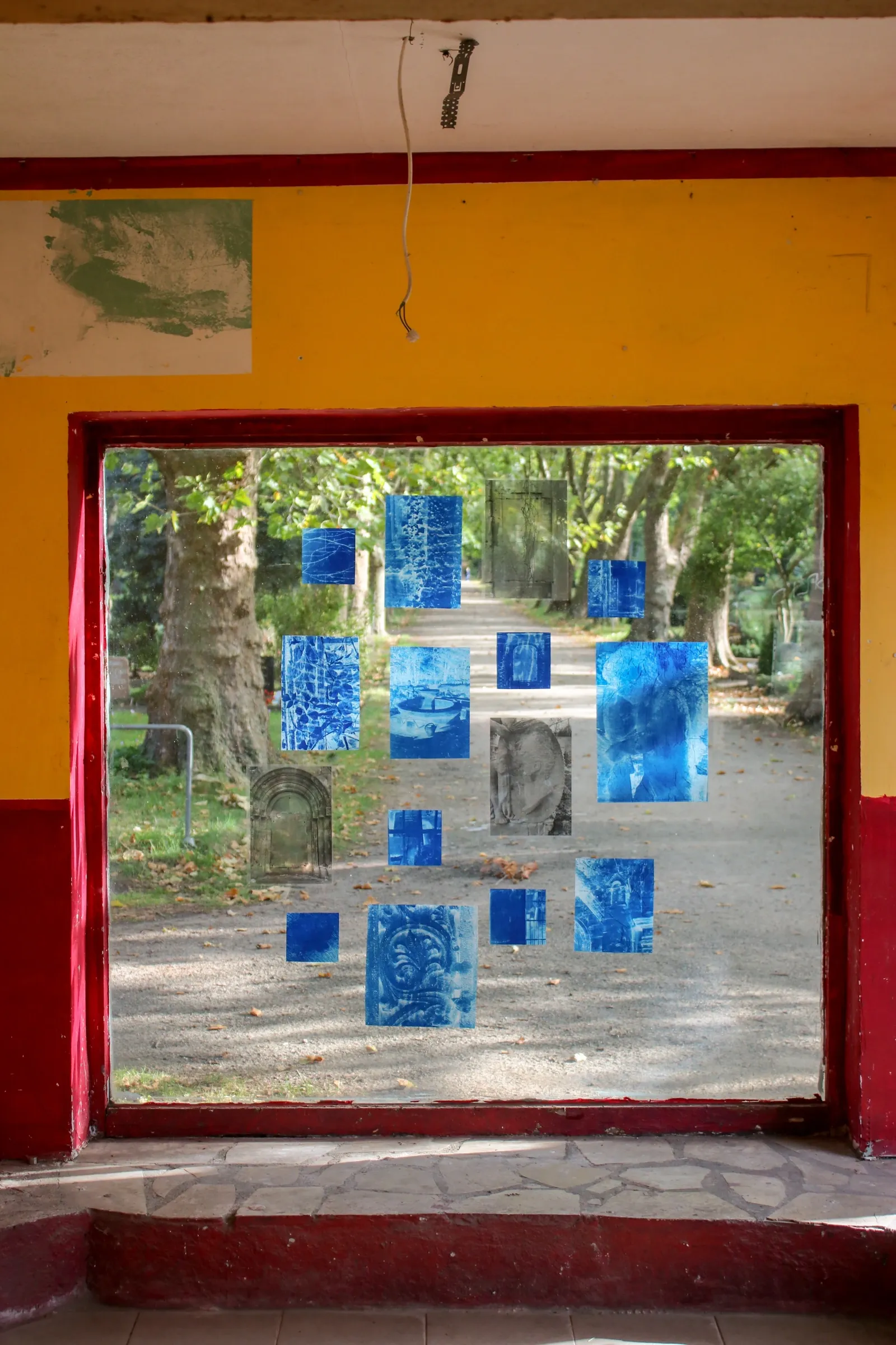 Aubrey&nbsp;Theobald&nbsp;and&nbsp;Kristina&nbsp;Popov,&nbsp;Phantom&nbsp;Pförtnerhaus&nbsp;Chapter&nbsp;3,&nbsp;Berlin,&nbsp;2026.&nbsp;Small&nbsp;blue&nbsp;and&nbsp;grey&nbsp;photographic&nbsp;prints&nbsp;on&nbsp;a&nbsp;window&nbsp;pane&nbsp;overlooking&nbsp;a&nbsp;cemetery&nbsp;path.