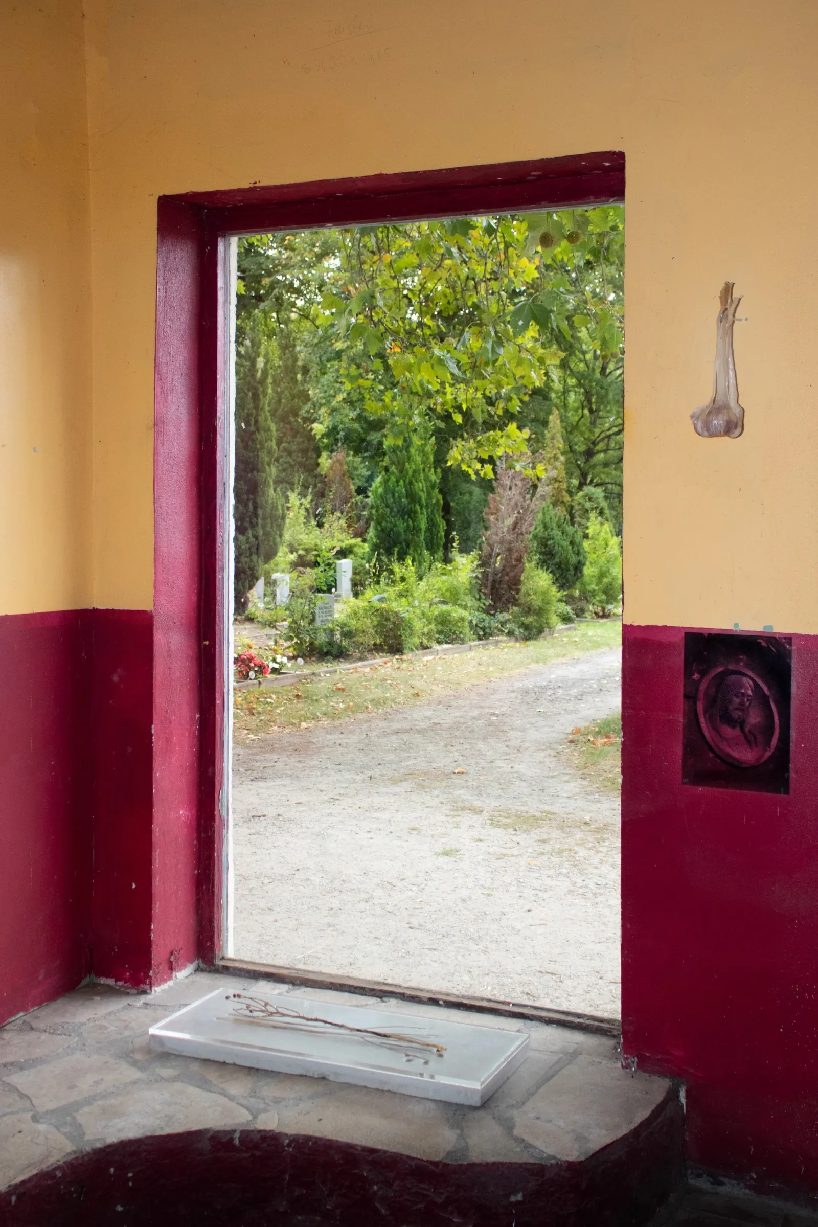 Aubrey&nbsp;Theobald&nbsp;and&nbsp;Kristina&nbsp;Popov,&nbsp;Phantom&nbsp;Pförtnerhaus&nbsp;Chapter&nbsp;3,&nbsp;Berlin,&nbsp;2026.&nbsp;Open&nbsp;doorway&nbsp;onto&nbsp;cemetery&nbsp;path,&nbsp;dried&nbsp;bulb&nbsp;on yellow&nbsp;wall,&nbsp;small&nbsp;relief&nbsp;print&nbsp;on&nbsp;crimson&nbsp;wall.
