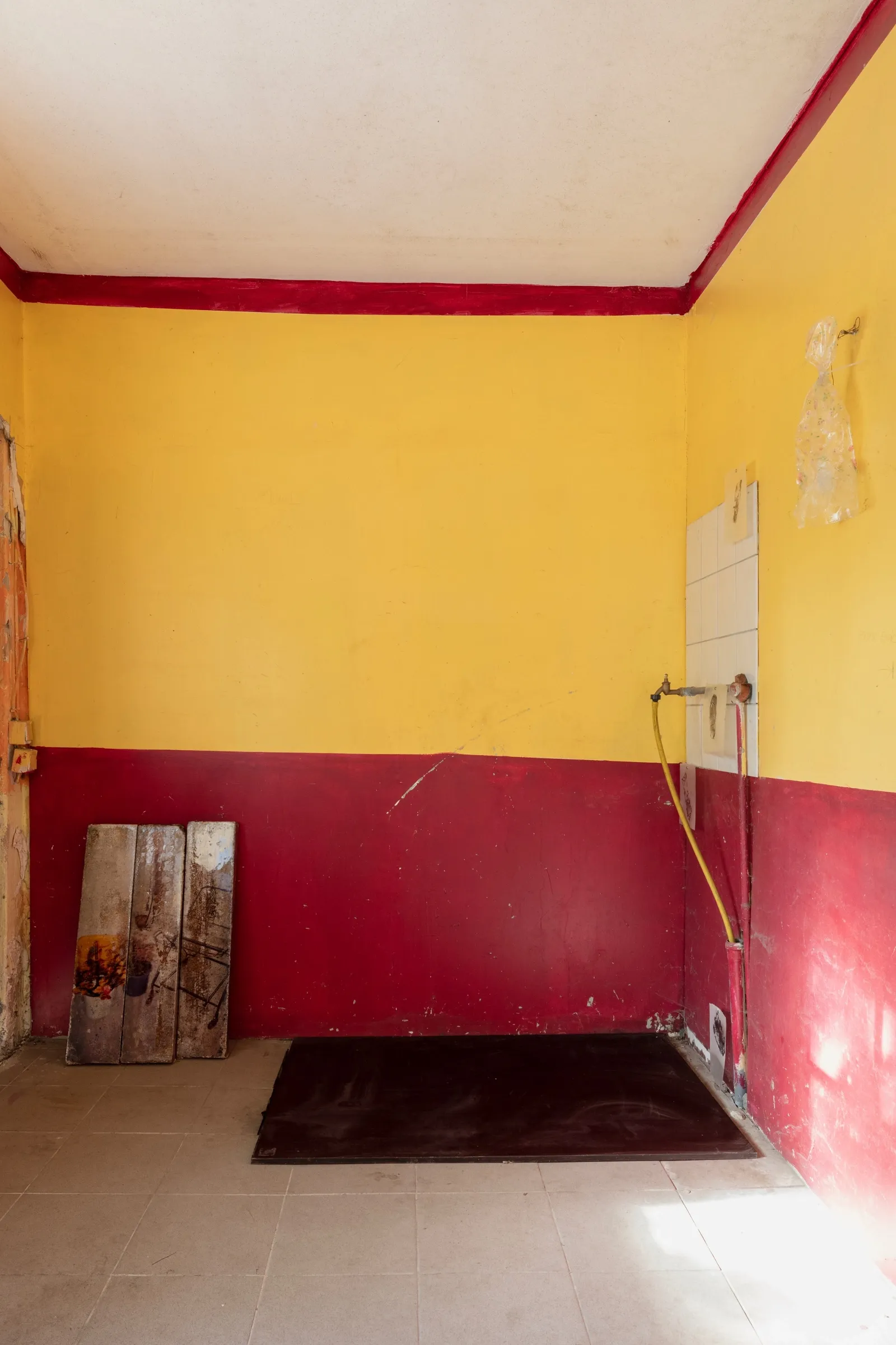 Aubrey&nbsp;Theobald&nbsp;and&nbsp;Kristina&nbsp;Popov,&nbsp;Phantom&nbsp;Pförtnerhaus. Yellow&nbsp;and&nbsp;crimson&nbsp;room&nbsp;with&nbsp;tiled&nbsp;wall&nbsp;section,&nbsp;yellow&nbsp;hose,&nbsp;leaning&nbsp;wooden&nbsp;panels&nbsp;and&nbsp;dark&nbsp;floor&nbsp;mat.