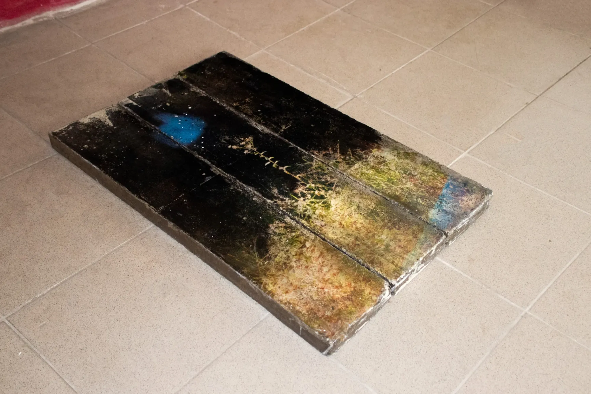 Aubrey&nbsp;Theobald&nbsp;and&nbsp;Kristina&nbsp;Popov,&nbsp;Phantom&nbsp;Pförtnerhaus&nbsp;Chapter&nbsp;3,&nbsp;Berlin,&nbsp;2026.&nbsp;Dark&nbsp;rectangular&nbsp;artwork&nbsp;with&nbsp;blue,&nbsp;gold&nbsp;and&nbsp;green&nbsp;markings&nbsp;lying&nbsp;on&nbsp;a&nbsp;beige&nbsp;tiled&nbsp;floor.