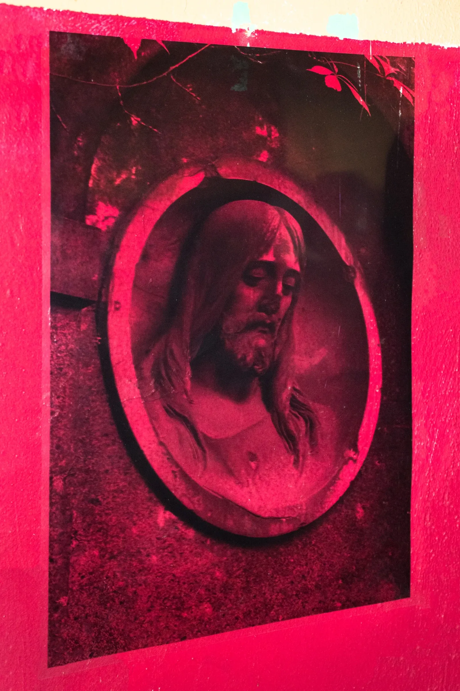 Aubrey&nbsp;Theobald&nbsp;and&nbsp;Kristina&nbsp;Popov,&nbsp;Phantom&nbsp;Pförtnerhaus&nbsp;Chapter&nbsp;3,&nbsp;Berlin,&nbsp;2026.&nbsp;Red-toned&nbsp;print&nbsp;of&nbsp;a&nbsp;bearded&nbsp;face&nbsp;in&nbsp;an&nbsp;oval&nbsp;cemetery&nbsp;medallion&nbsp;mounted&nbsp;on&nbsp;a&nbsp;crimson&nbsp;wall.