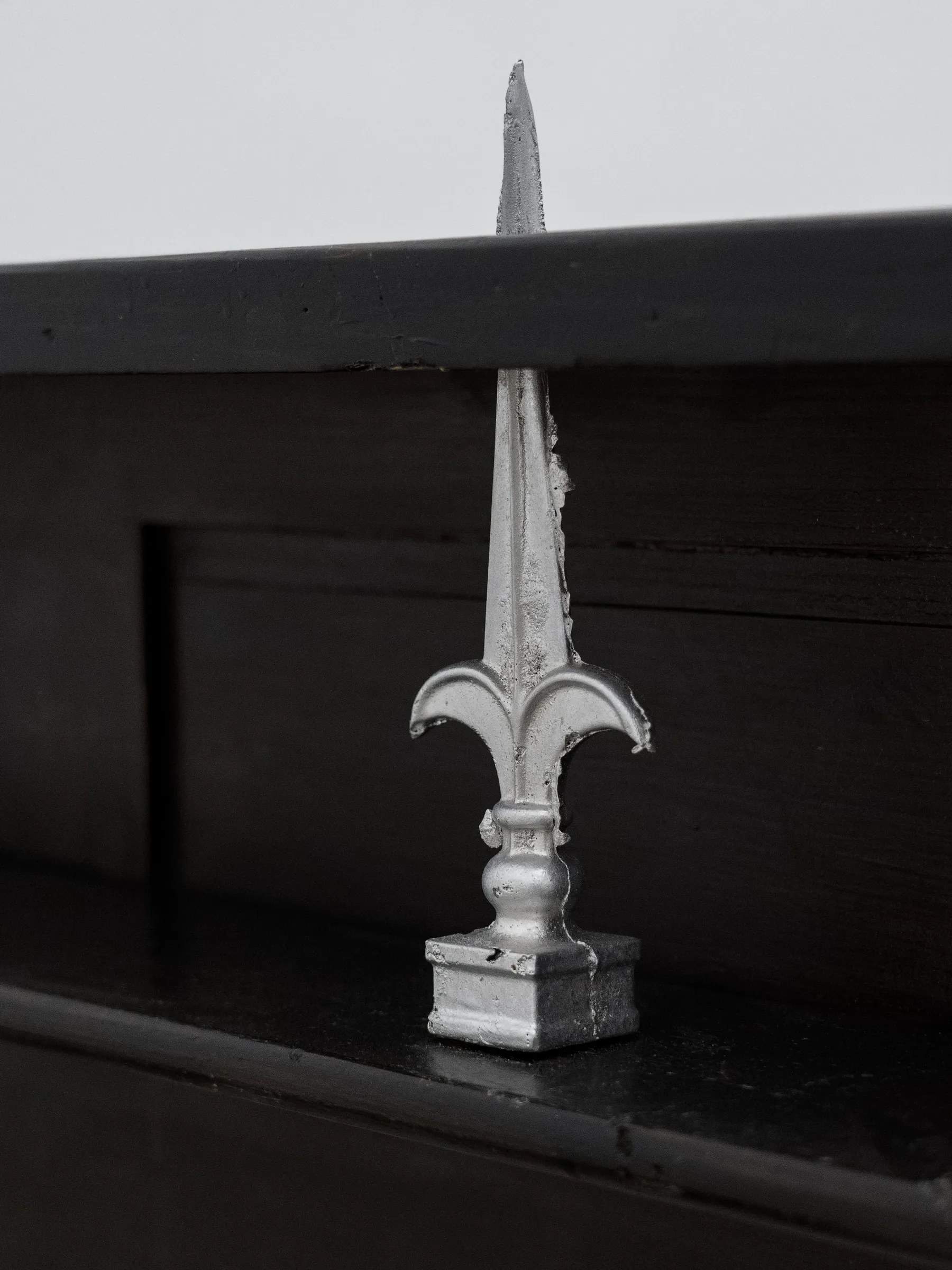 Exhibition review of AUSGEWEIDET / GUTTED at WAF Galerie, Vienna, 2026. Silver fleur-de-lis spike with a sharp tip on the black wooden pew.