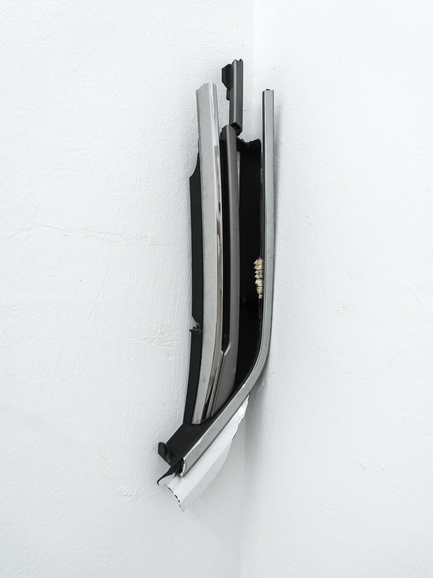 Exhibition review of AUSGEWEIDET / GUTTED at WAF Galerie, Vienna, 2026. Chrome and black car trim bent into a white wall corner, a row of teeth in the gap.