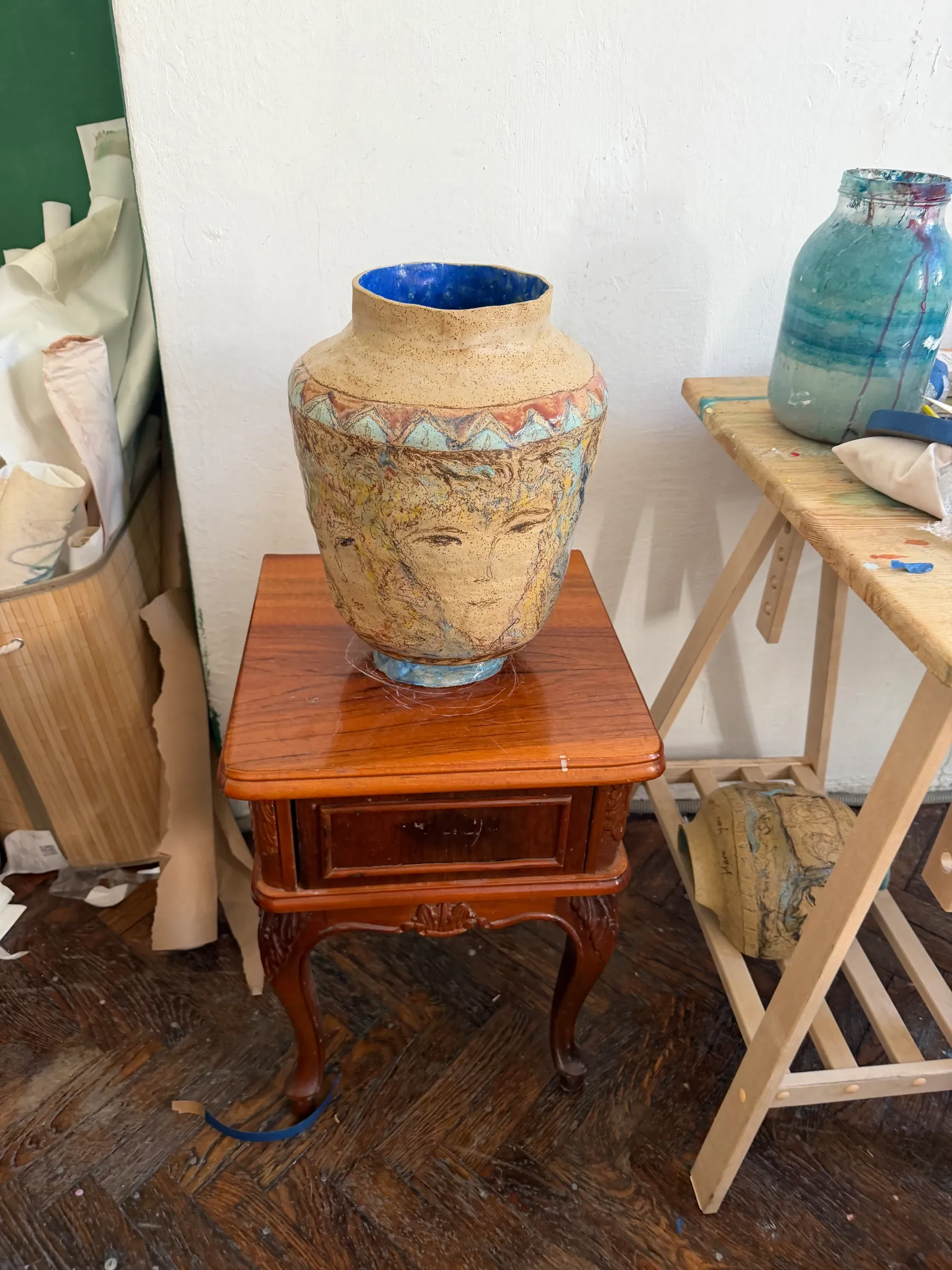 Christina Arark: ceramic vase with a drawn face on a wooden side table in the studio, trestle and canvases behind.