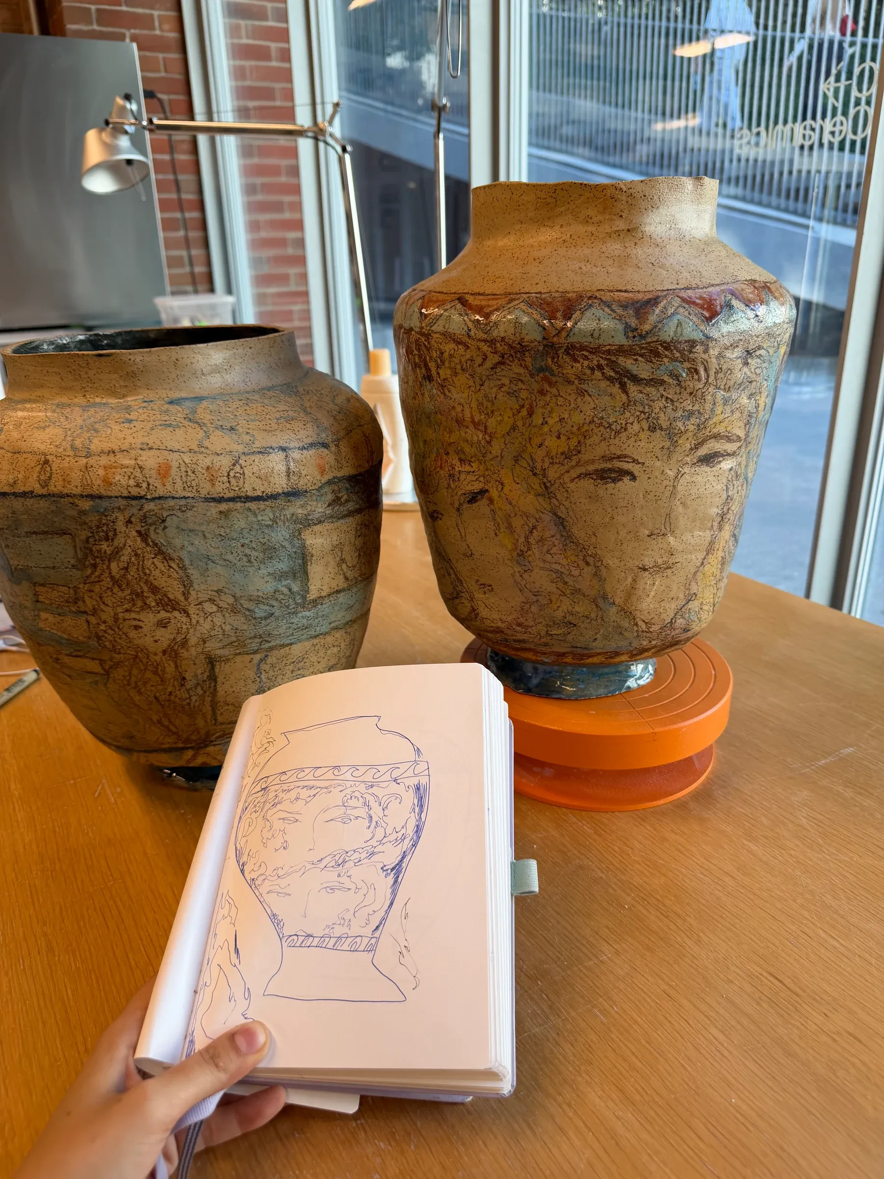 ALT: Christina Arark: open sketchbook with a blue line drawing of a vase beside two ceramic vases on a wooden table.