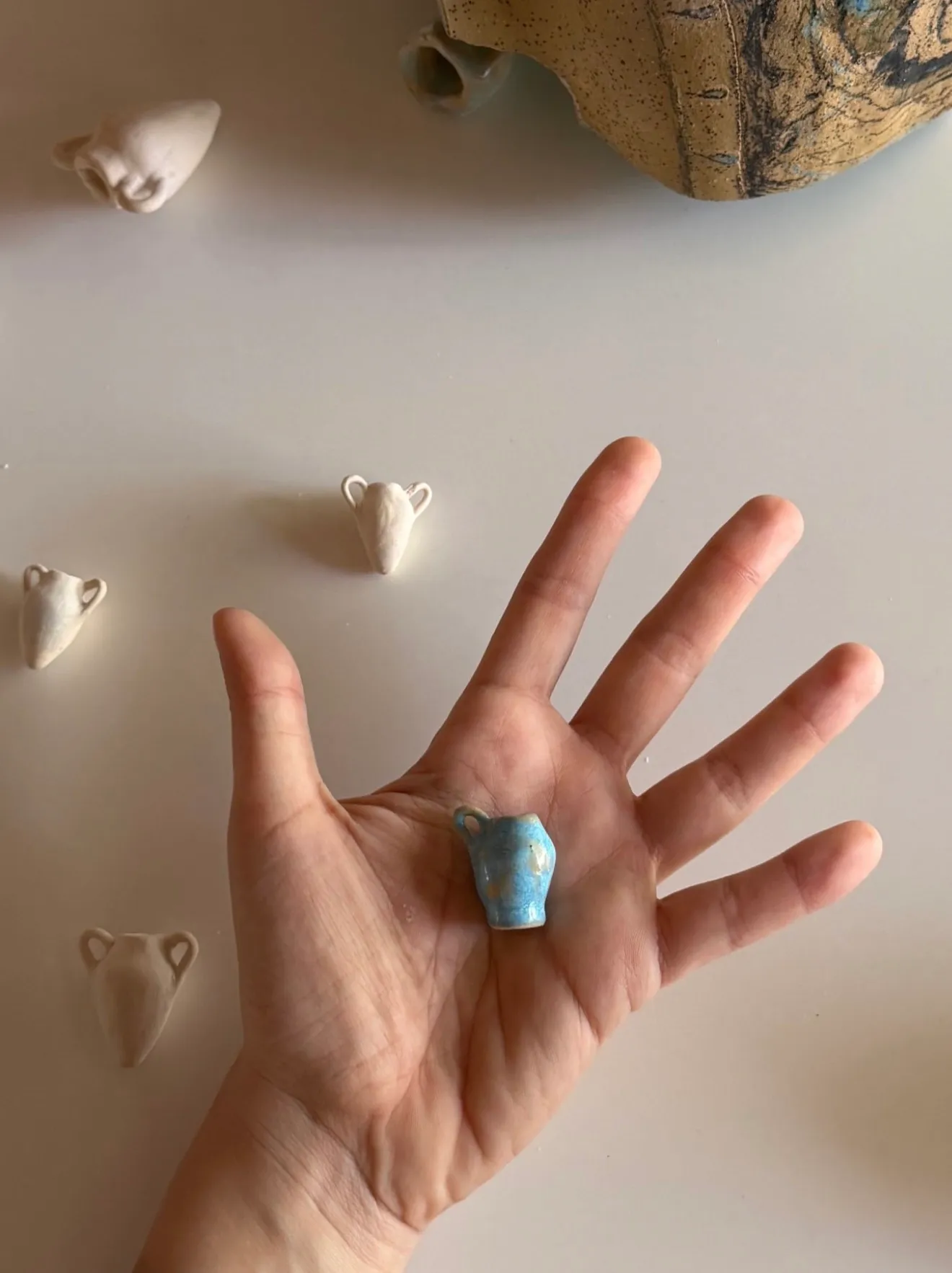 ALT: Christina Arark: a hand holding a small blue ceramic vessel, small white vessels on the surface around it.