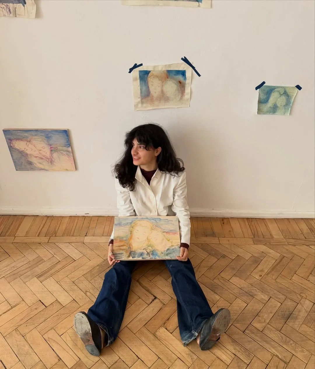 ALT: Christina Arark: artist sitting on a wooden floor holding a small painting, more paintings taped to the wall behind her.