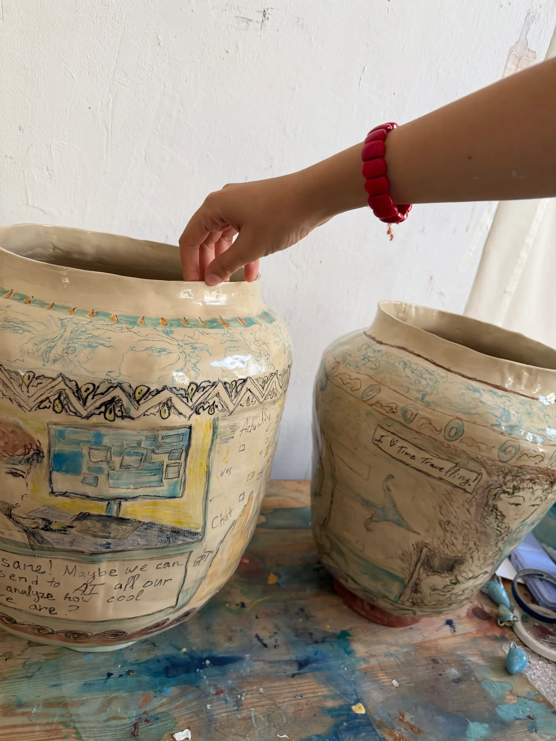 ALT: Christina Arark: two ceramic vases with drawn text boxes on a paint-stained table, a hand with a red bracelet on the rim.