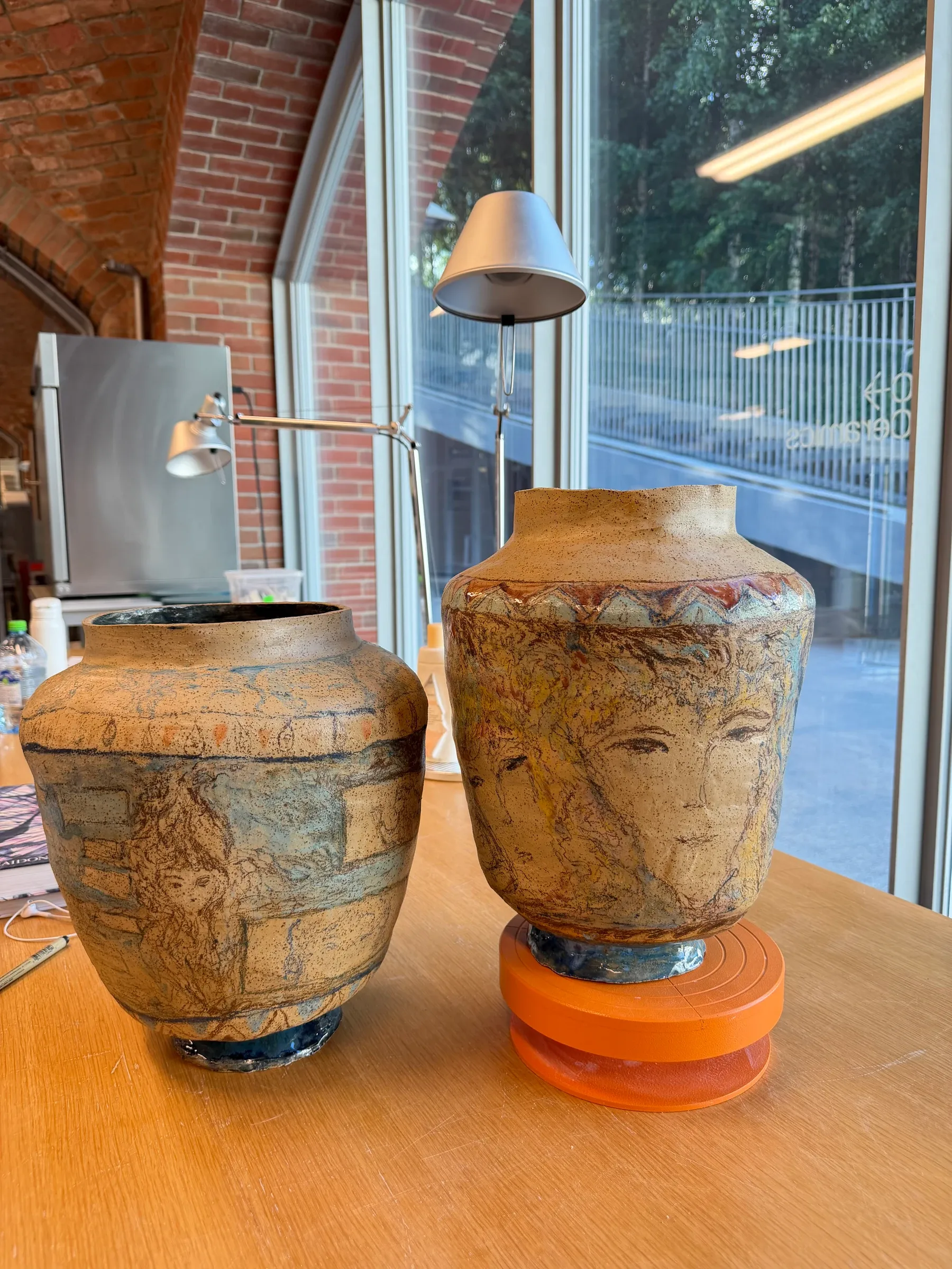 ALT: Christina Arark: two speckled ceramic vases with drawn faces on a wooden table by a window, one on an orange stand.