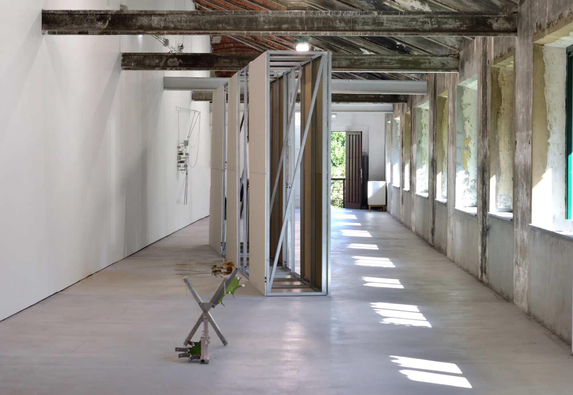 Exhibition review of Gabriel Alonso: A Long Chemical Dream at ATM Galería, Gijón, 2026. Metal X-shaped floor work before the green printed wall, daylight at the far end.
