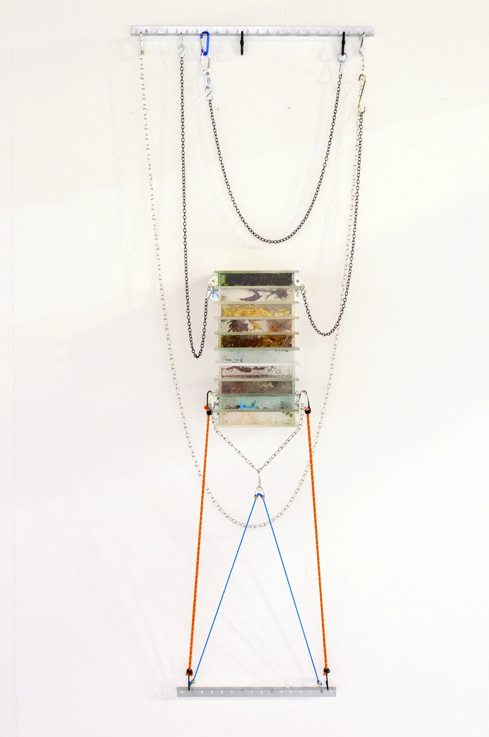 Exhibition review of Gabriel Alonso: A Long Chemical Dream at ATM Galería, Gijón, 2026. Stacked acrylic trays of plant matter hung on chains with orange and blue cords.