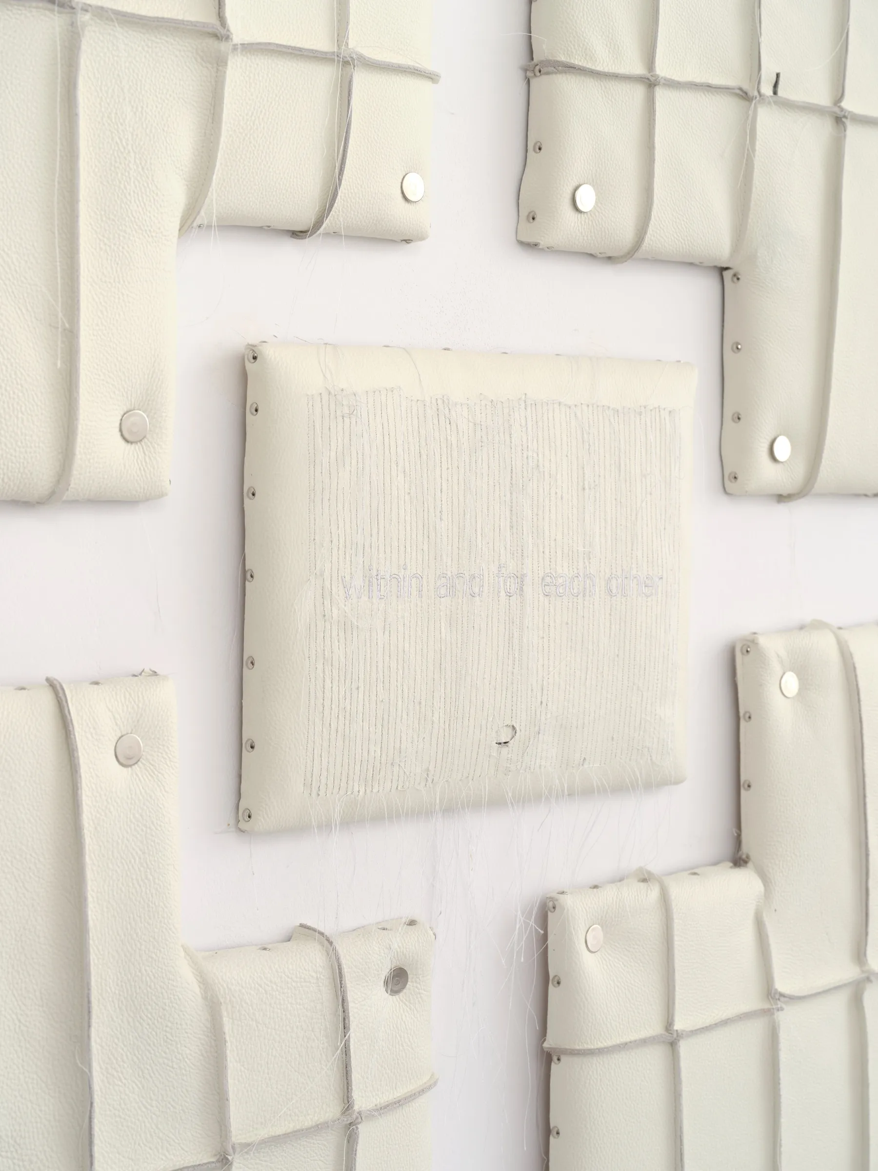 Close-up of cream padded sculptural panels with metal studs surrounding a central plate bearing faint lettering. Jan Baszak, Within and For Each Other, WHOISPOLA, Warsaw, 2026.