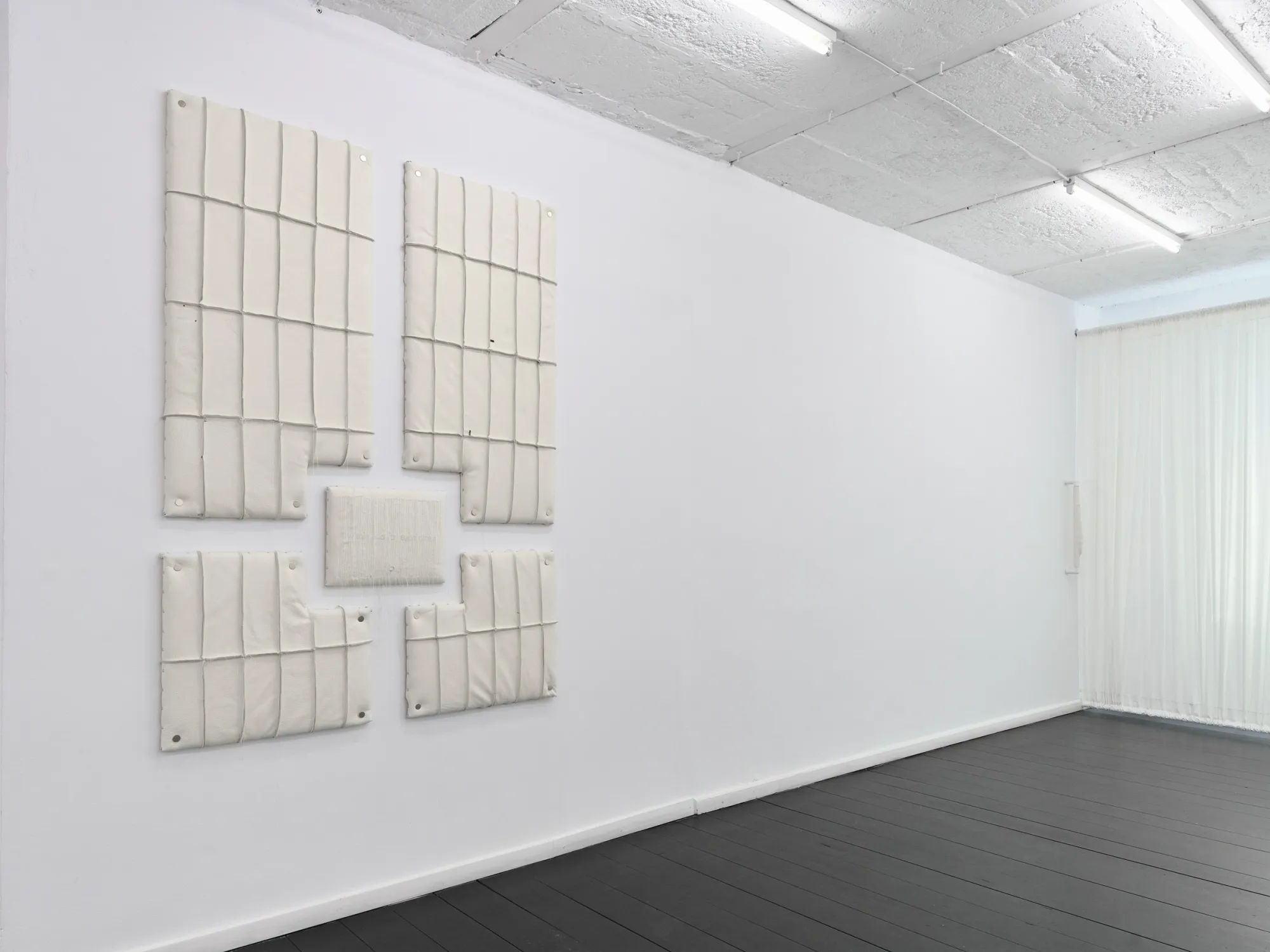 Cream padded panels installed on a white gallery wall beside floor-length sheer curtains. Jan Baszak, Within and For Each Other, Warsaw, 2026.