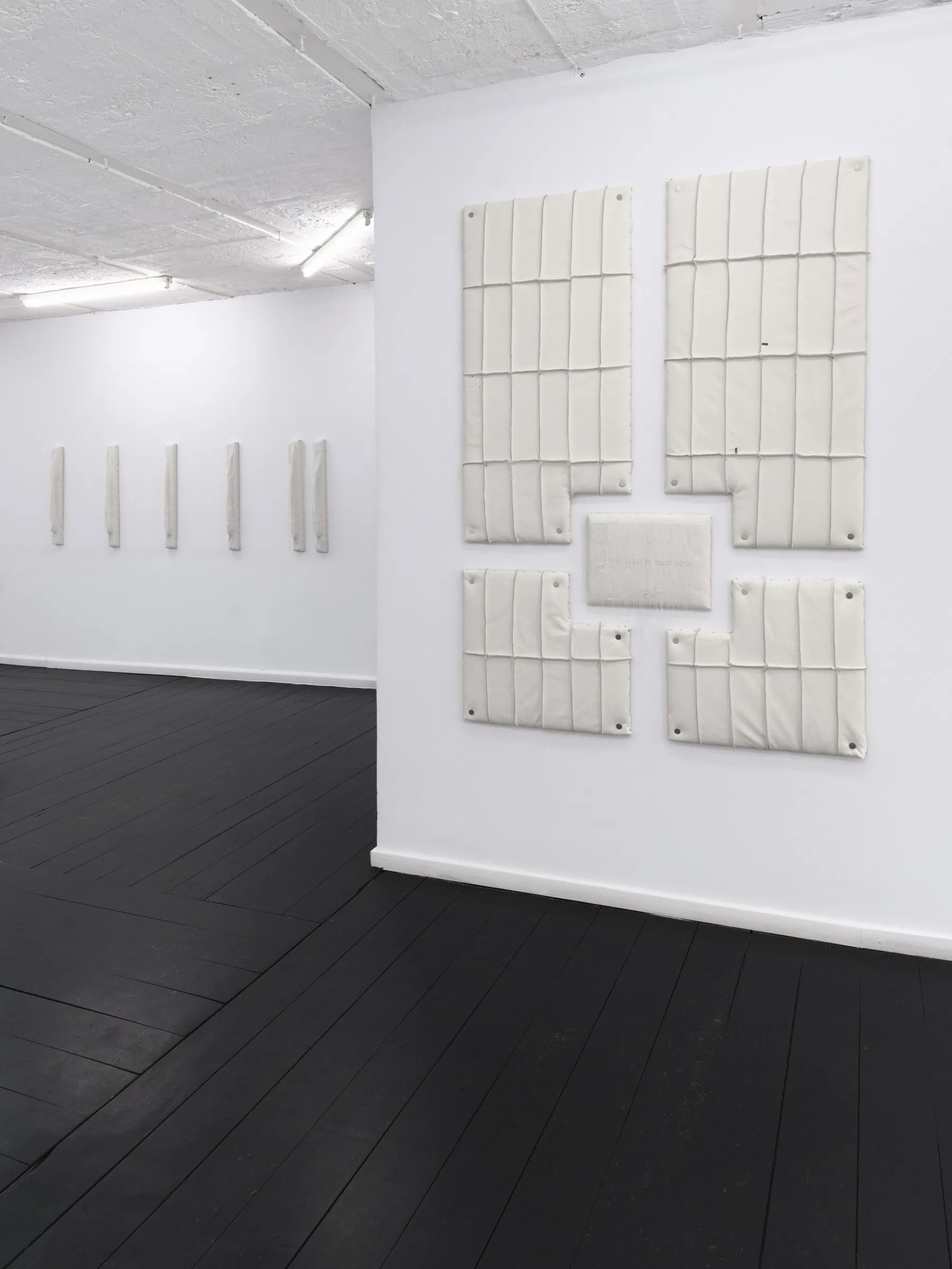 Installation view of cream padded wall panels in the foreground, with narrow vertical sculptures visible on the opposite gallery wall. Jan Baszak, WHOISPOLA, Warsaw, 2026.