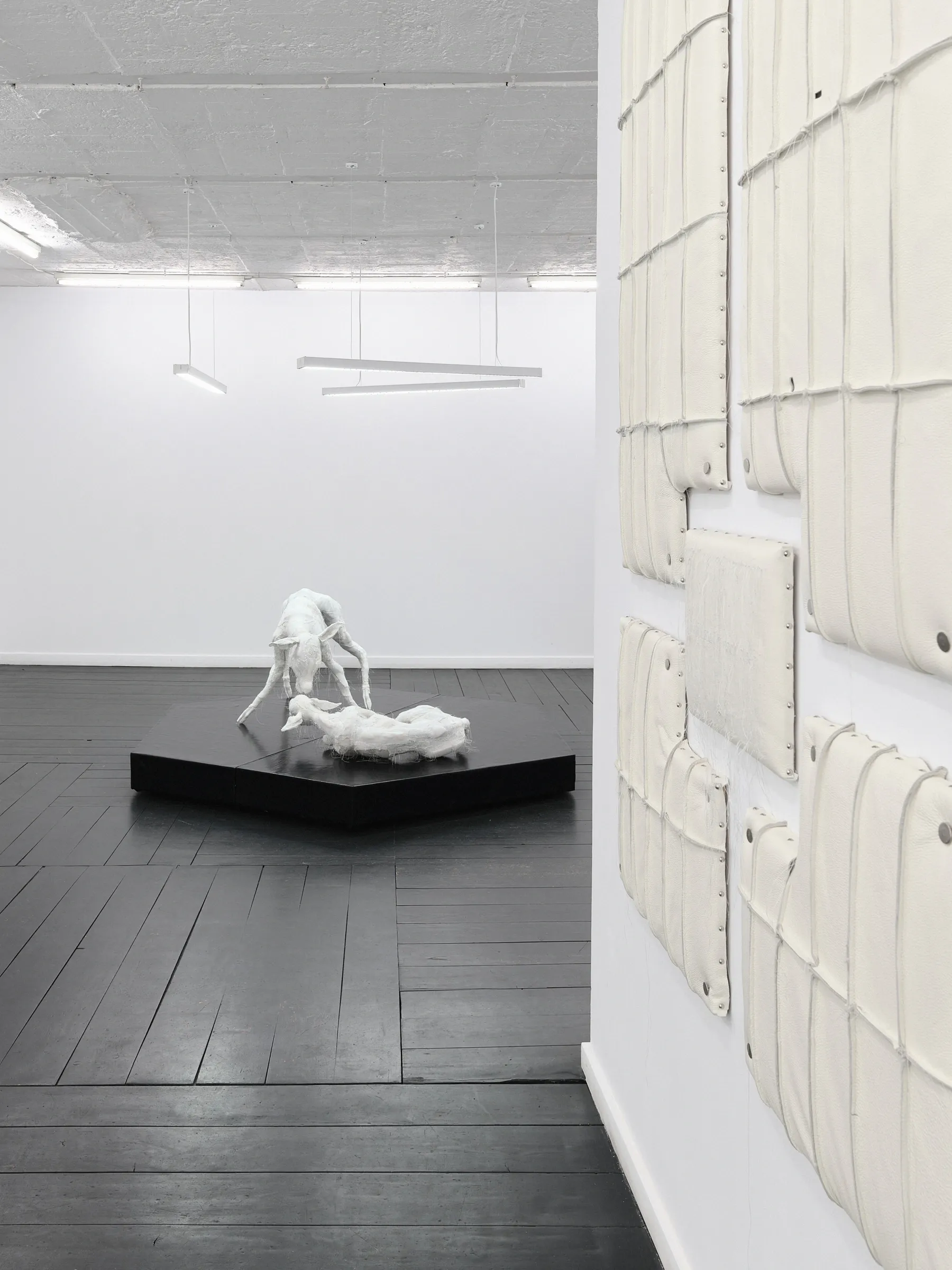 White deer sculptures on a black plinth viewed through the gallery past a wall of cream padded panels. Jan Baszak, Within and For Each Other, WHOISPOLA, Warsaw, 2026.