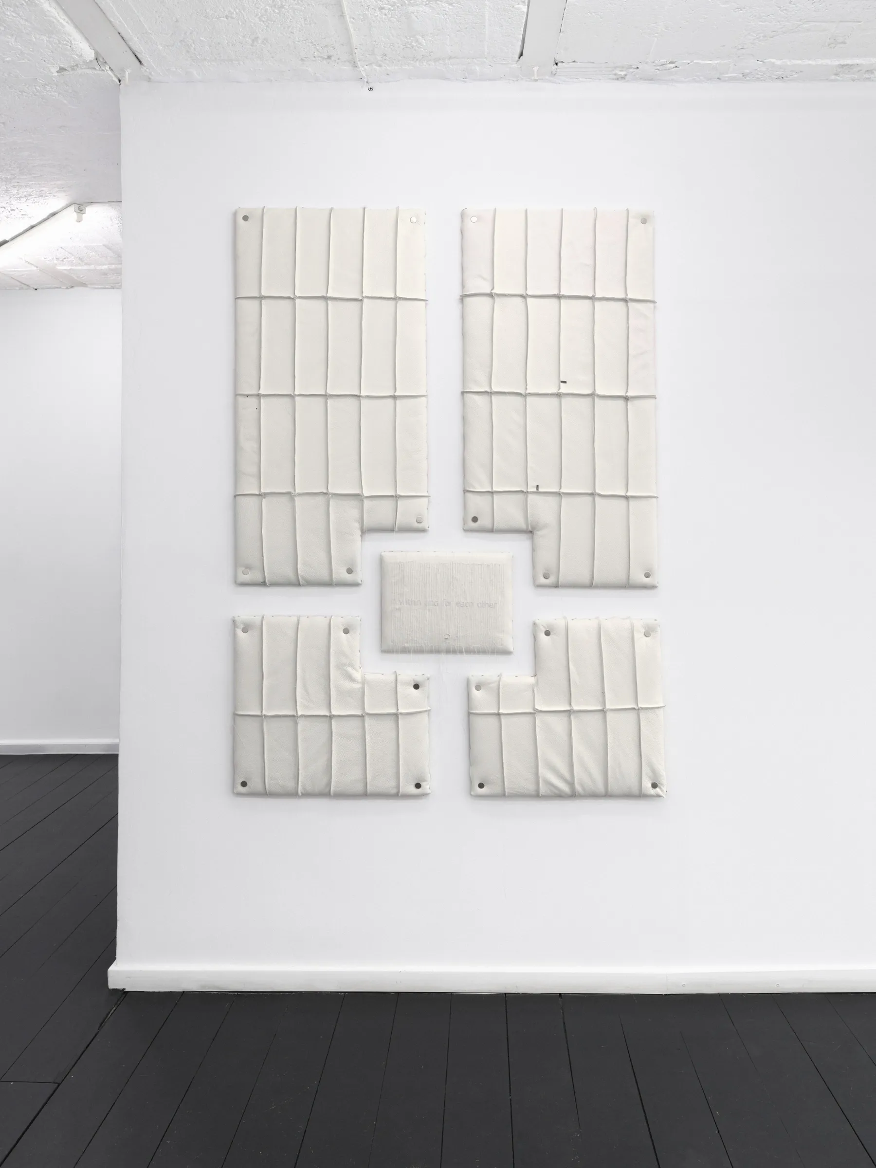 Cream quilted wall panels with metal studs arranged around a central rectangle. Jan Baszak, Within and For Each Other, WHOISPOLA, Warsaw, 2026.