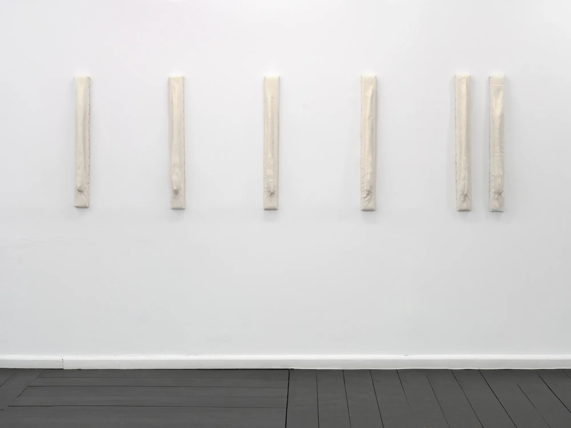 Six narrow upright cream padded bars displayed in a row on a white gallery wall. Jan Baszak, Within and For Each Other, WHOISPOLA, Warsaw, 2026.