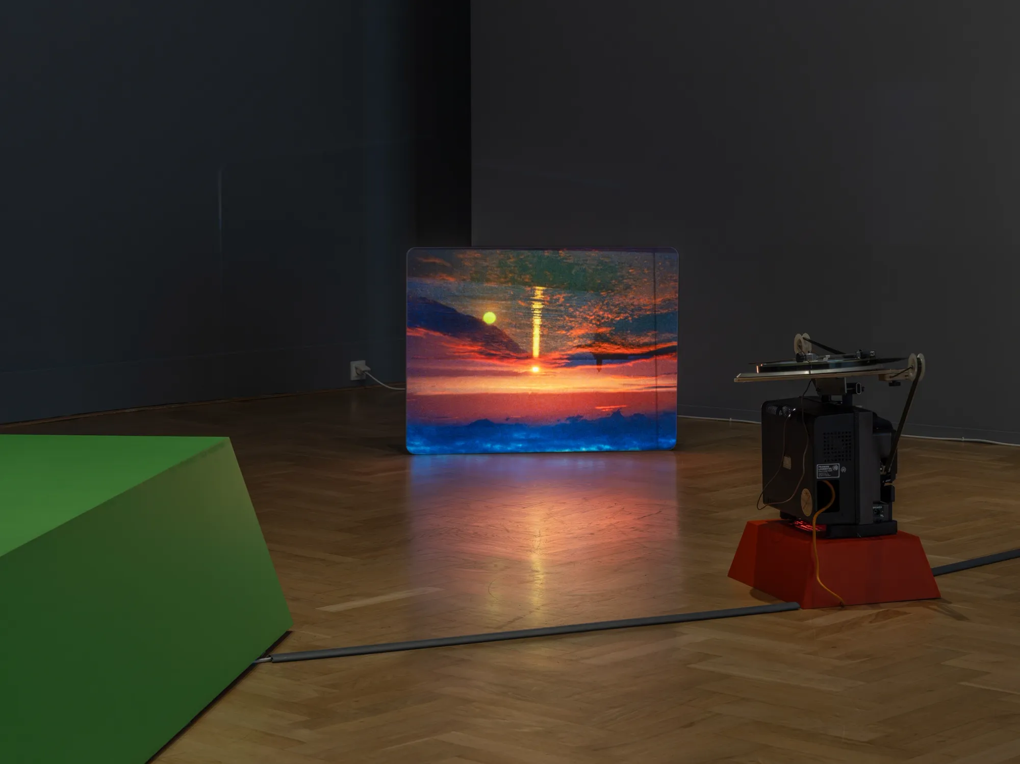 ALT: João Maria Gusmão, Zachęta, Warsaw: projected red sunset over a blue horizon beside a 16mm projector on a red base, green plinth at left.