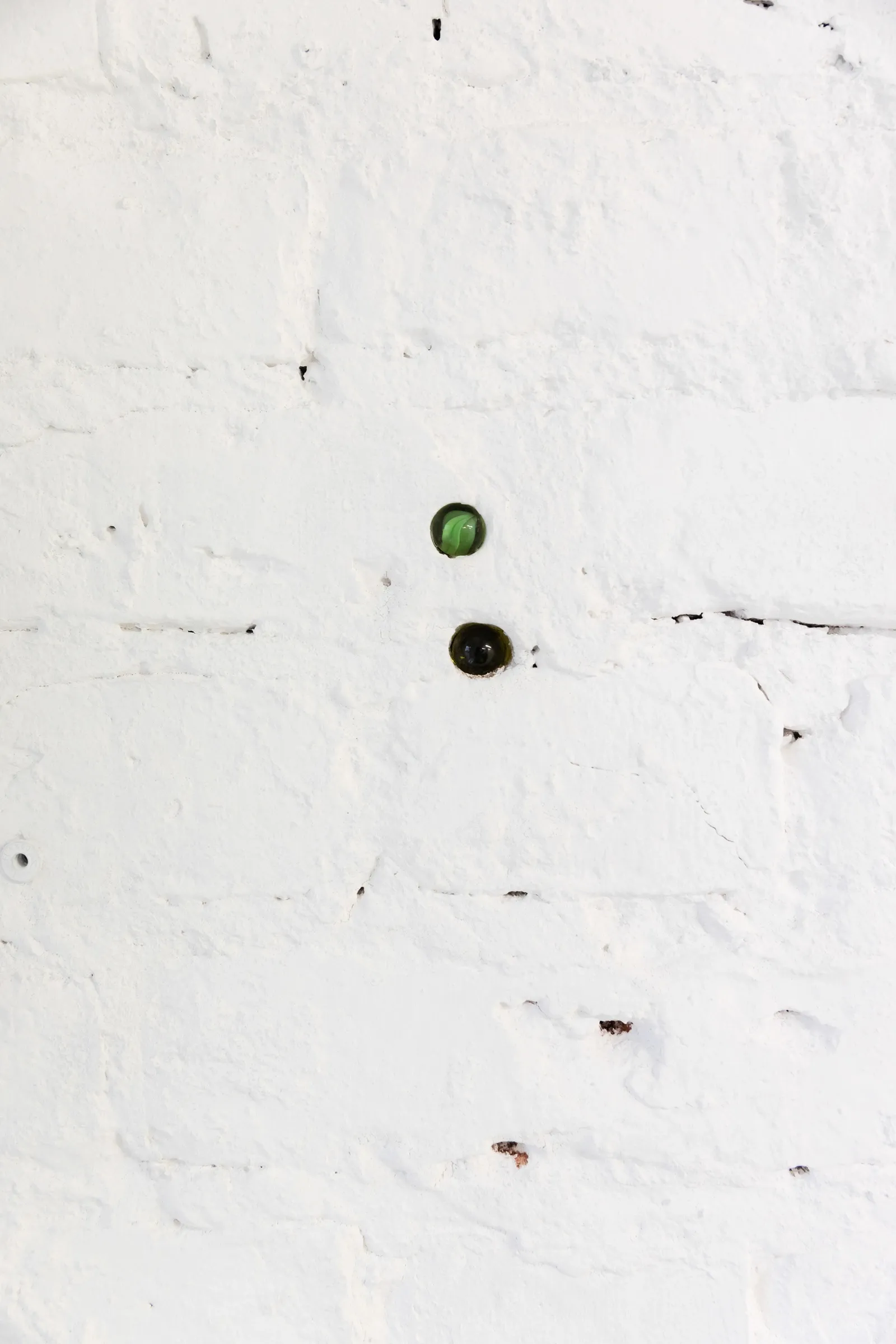 Exhibition review of Krys Huba: falling into you at soft power, Berlin, 2026. Two green glass marbles set into a white-painted brick wall.
